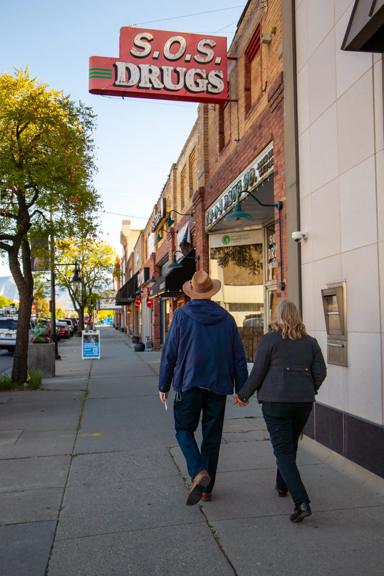 Two people walking hand-in-hand down a sidewalk in a small town. The man is wearing a blue jacket and a wide-brimmed hat, while the woman is in a dark jacket. They are walking past storefronts, one of which is a drugstore with a sign that says 'S.O.S. Drugs.' There are trees with green leaves and parked cars along the street.