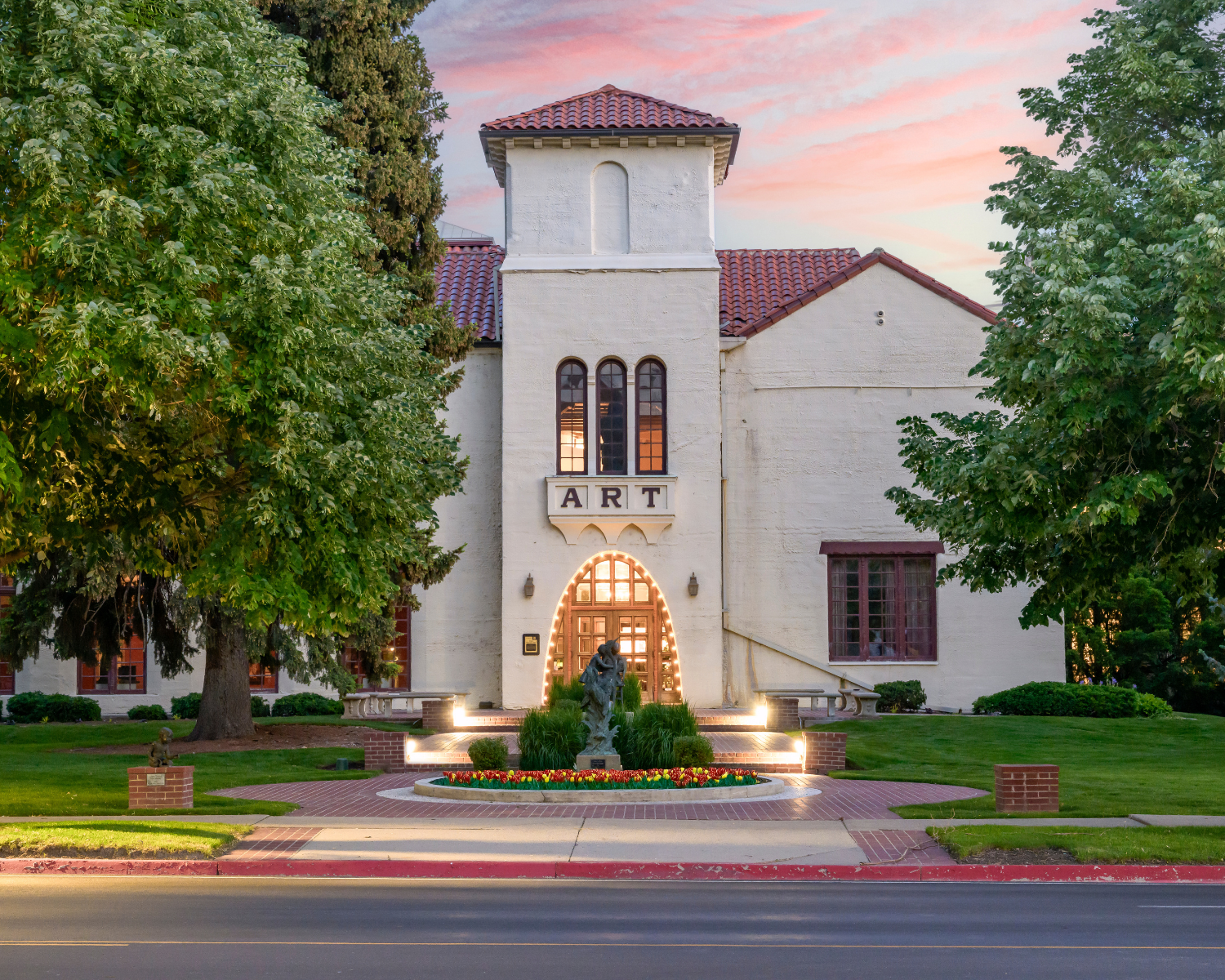 Springville Museum of Art building with a sign that reads 'ART' above the entrance, flanked by trees at sunset, featuring a symmetrical facade, arched doorway with lights, and a statue in front surrounded by flower beds.