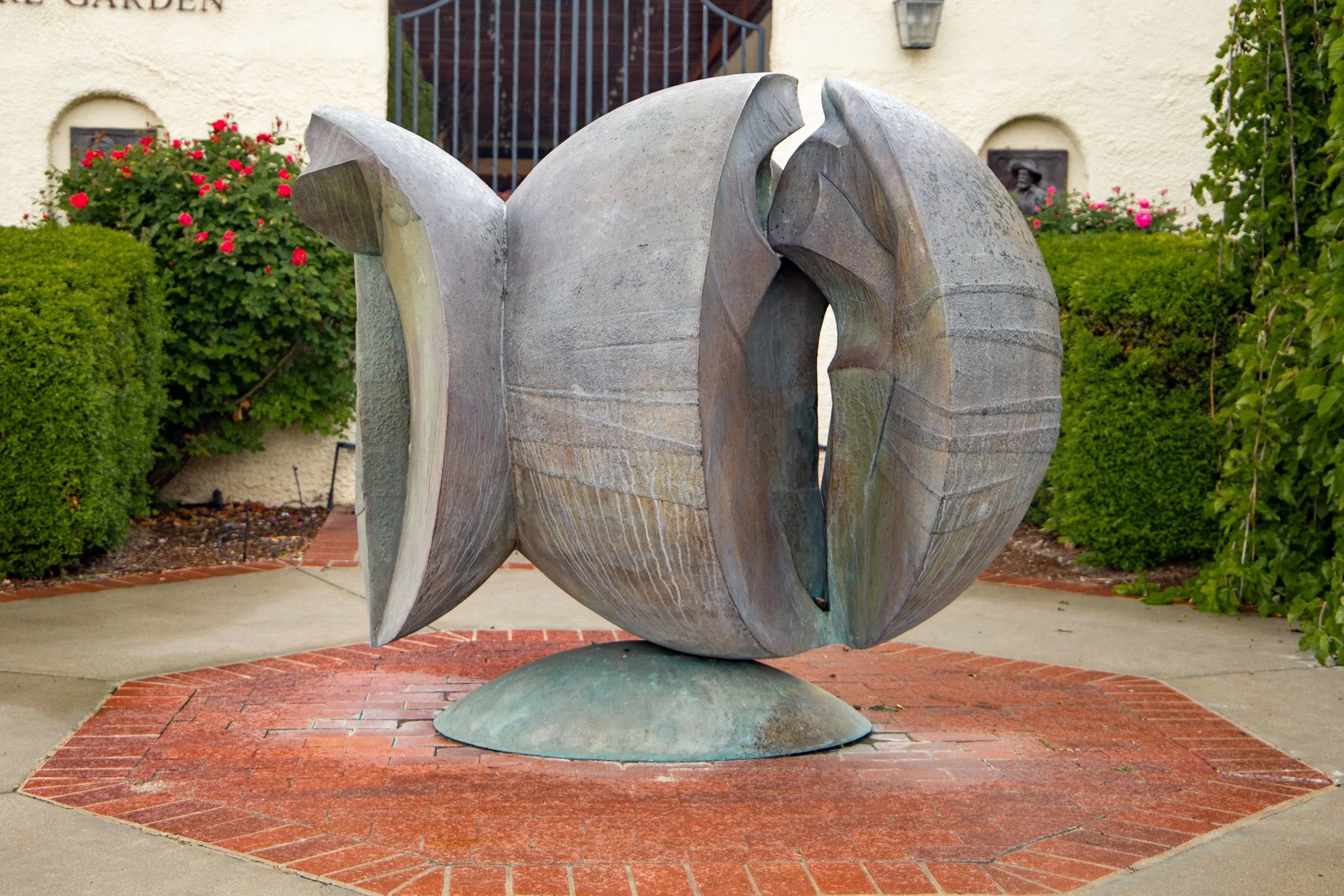 A modern abstract metal sculpture resembling an animal (possibly a horse or dog) situated on a brick and concrete paved area in front of a building with a staircase, surrounded by landscaped bushes and flowers.