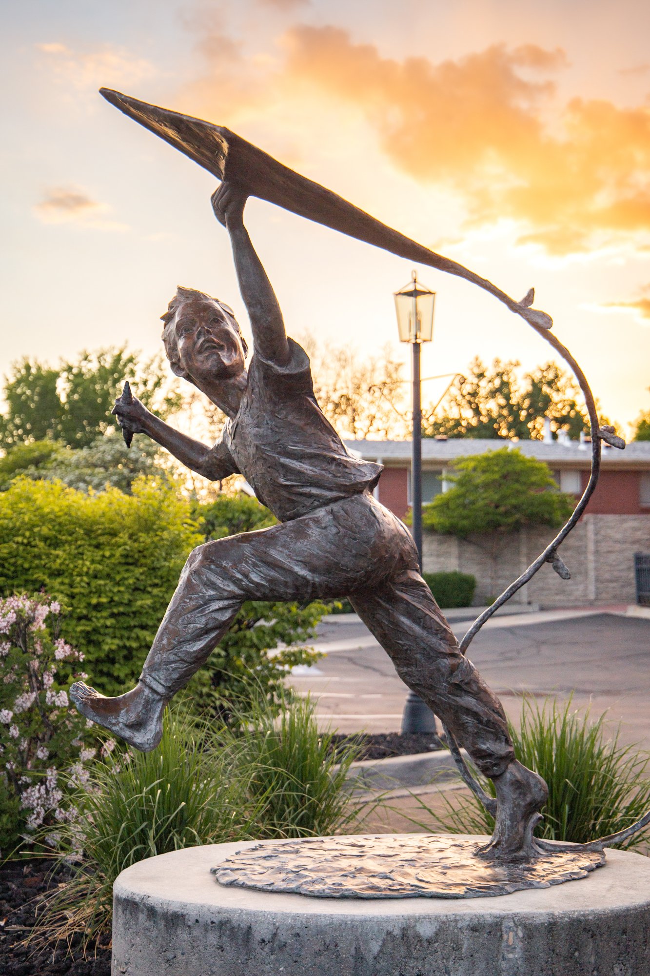 Bronze statue of a young boy running and leaping with a fishing pole in one hand, set on a circular stone pedestal outdoors at sunset with clouds, trees, and a street lamp in the background.