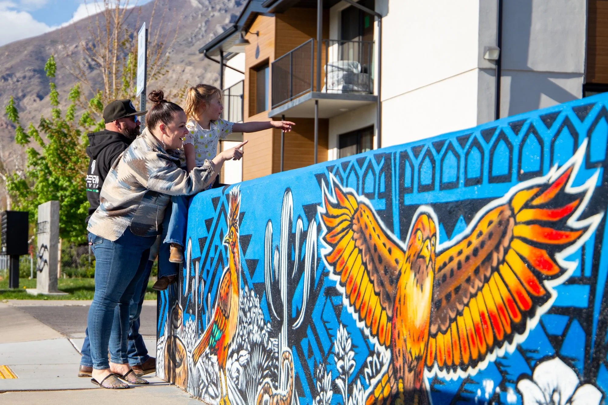 Three people, two women and one man, are standing by a colorful painted fence, with a young girl sitting on the woman's shoulders, pointing at a building. The background features a mountain, trees, and a partly cloudy sky.