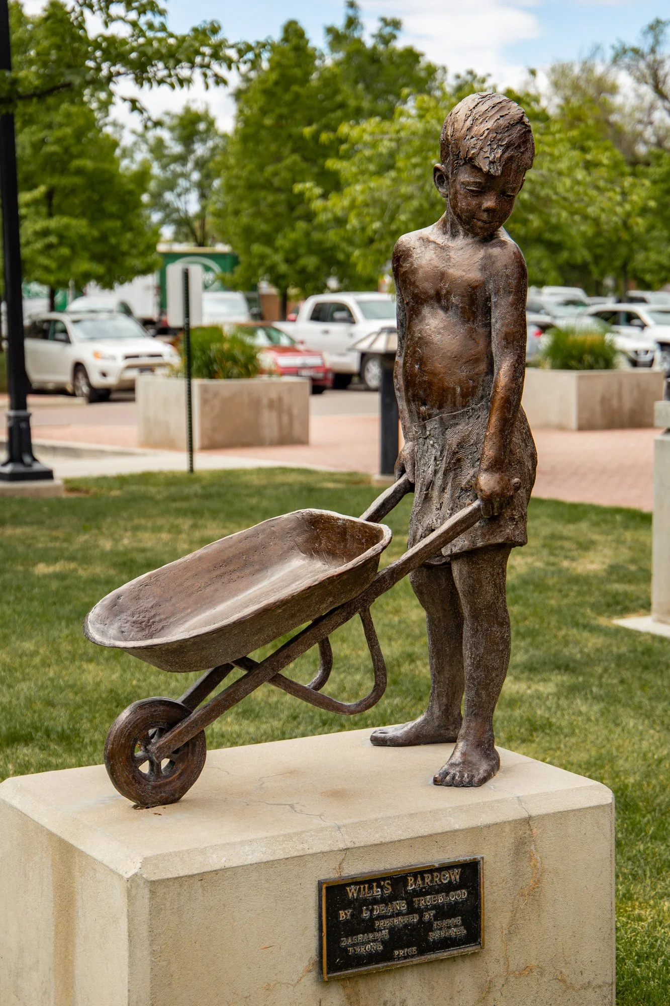 A bronze statue of a young boy pushing a wheelbarrow, standing on a stone pedestal in a park with trees and parked cars in the background.