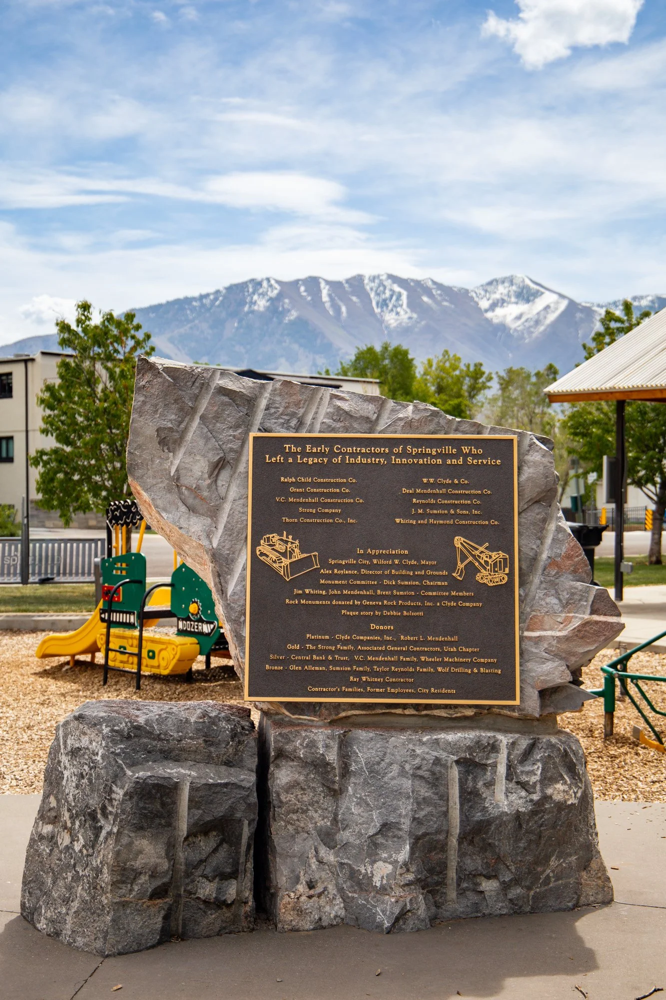 A commemorative plaque on a large rock monument in a park, with snow-capped mountains and a partly cloudy sky in the background. The plaque honors early contractors of Springville, Utah.