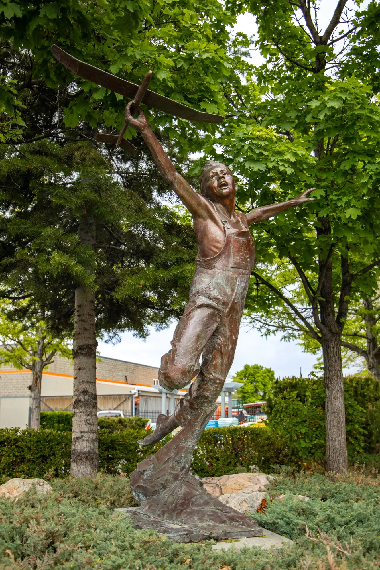 Bronze memorial statue of a young boy in mid-flight with arms outstretched, holding a large airplane overhead, amidst green trees and shrubs.