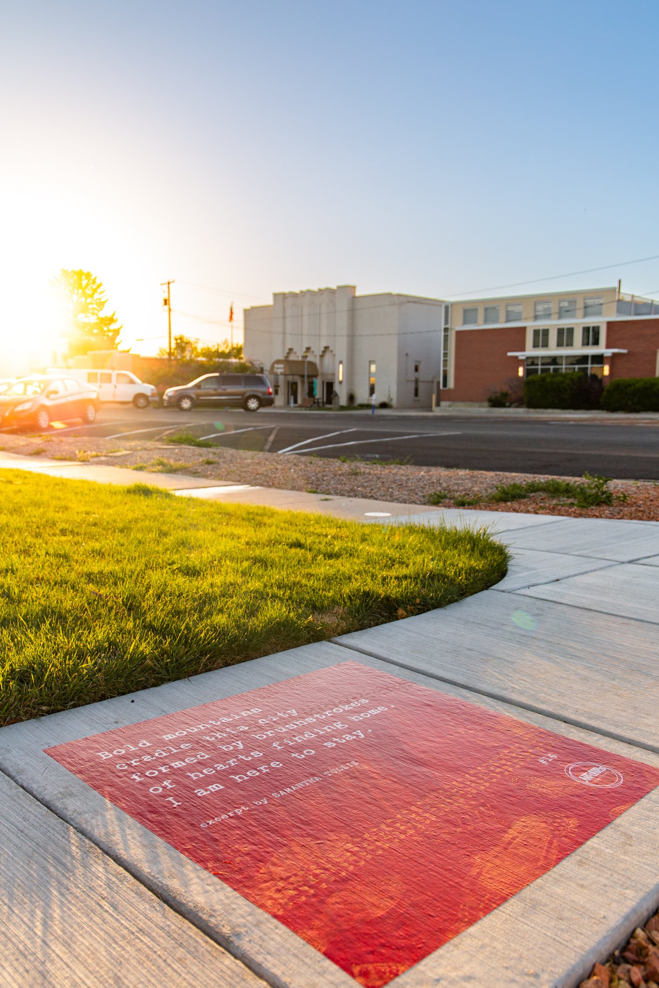 Red sidewalk poem installed at a curved sidewalk corner near the Senior Center building in Springville, photographed in warm evening light with surrounding lawn and streetscape.
