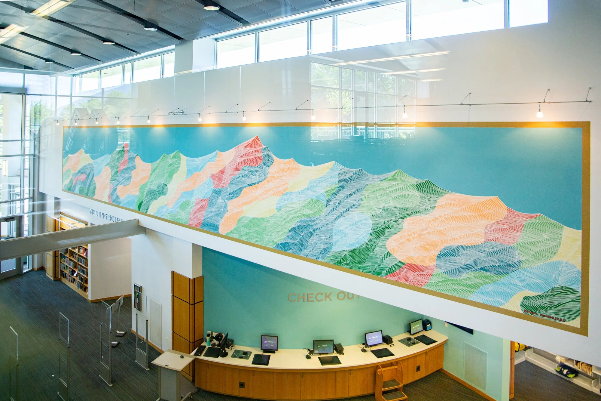 Interior view of a modern library or public space with a large colorful mountain-themed mural on the wall, a check-out counter with computers, and multiple levels with bookshelves and seating areas, illuminated by natural light from large windows.