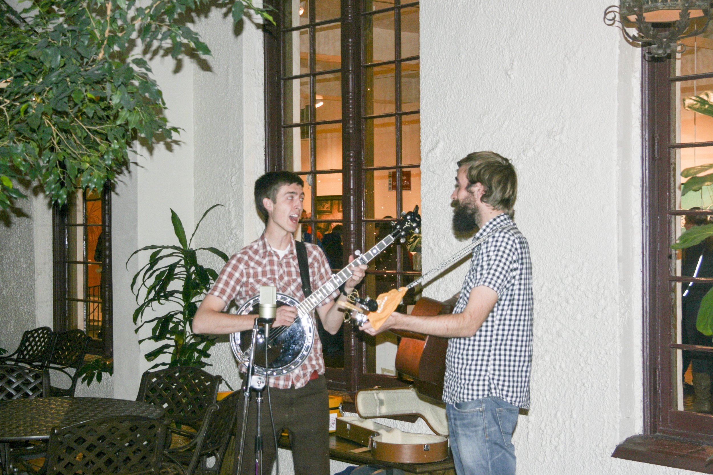 Two men are playing guitars and singing together indoors near large windows, with plants and furniture around them.