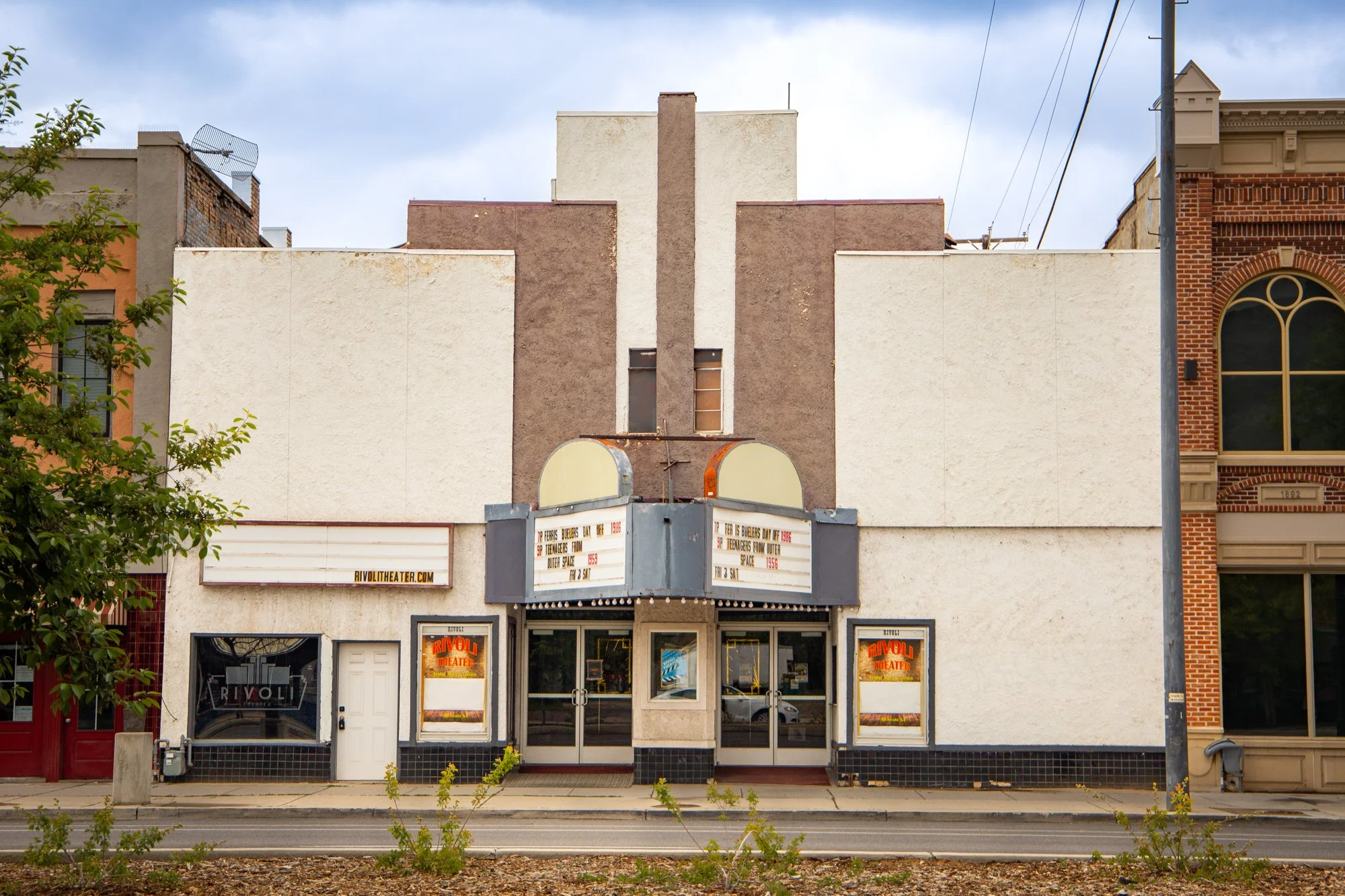 An old theater building with a white facade, brown vertical accents, and an marquee sign. The marquee has some text and is surrounded by large windows displaying posters.