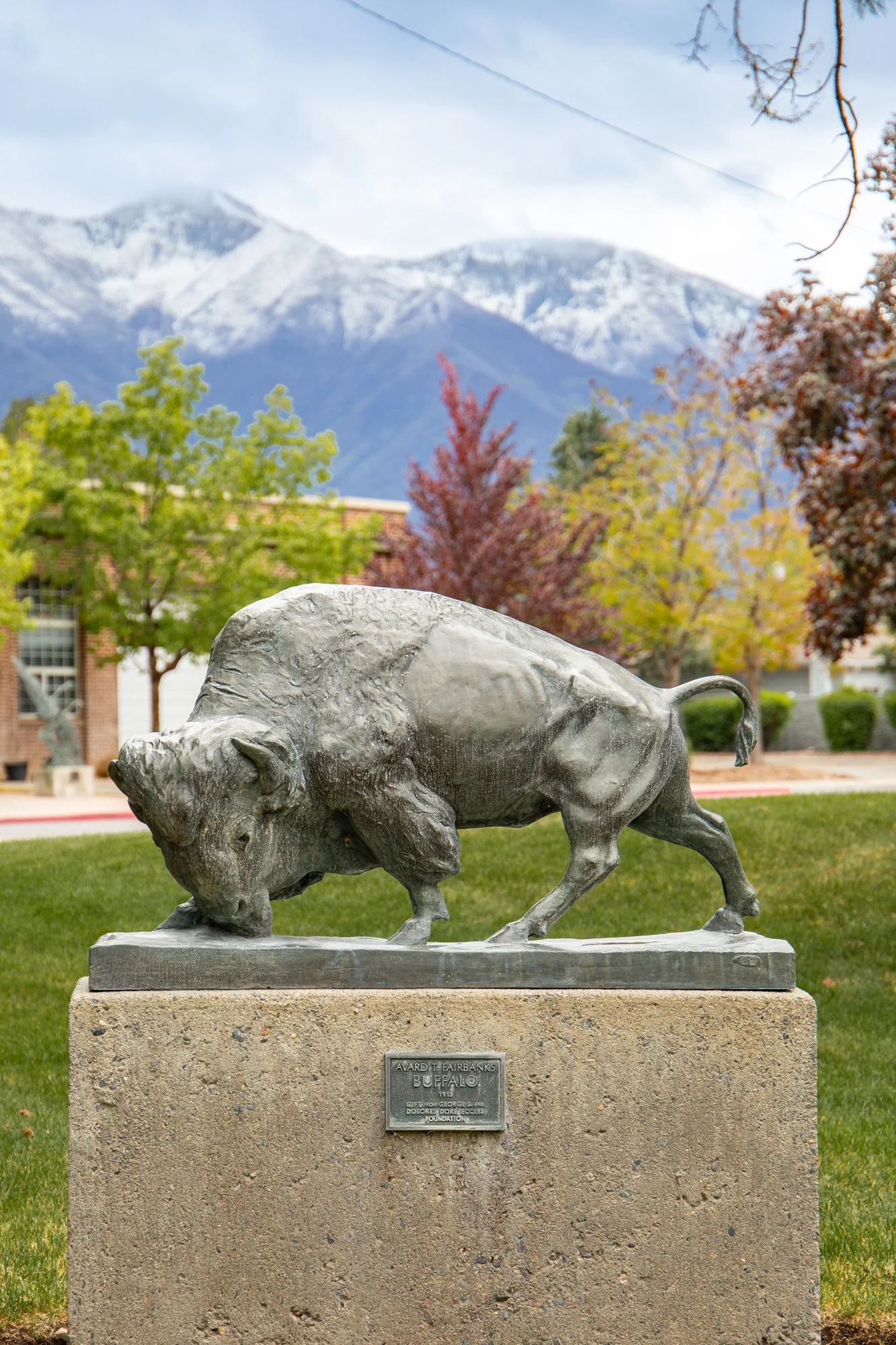 Bronze statue of a bison on a stone pedestal with a mountain range and trees in the background.