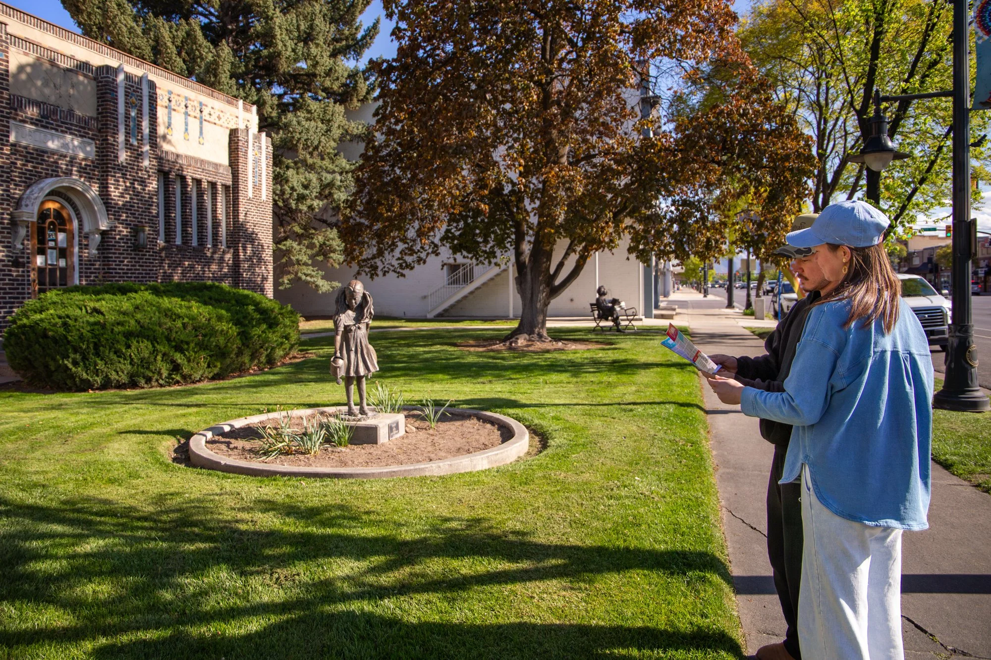 A couple with hats reading brochures stand on a sidewalk in front of a park with trees, a statue of a young girl, and buildings in the background.