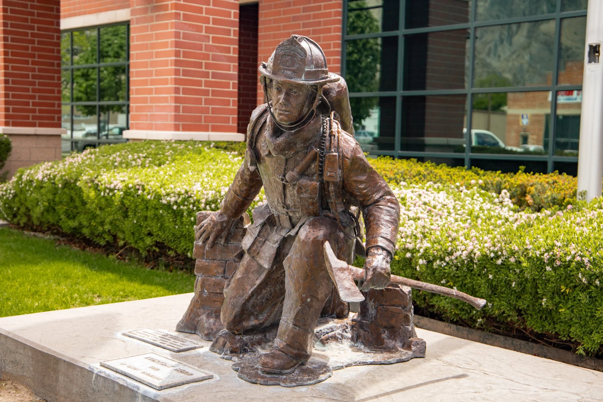 Bronze statue of a firefighter with a helmet, kneeling and holding a tool, set on a stone foundation with plaques, surrounded by green bushes and flowers in front of a brick and glass building.