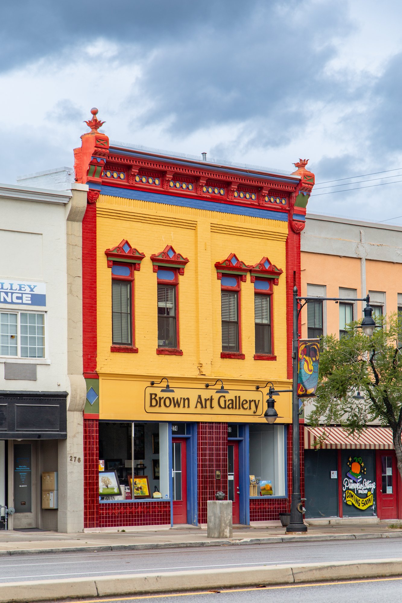 Colorful building with a yellow facade, red trim, and blue accents, housing the Brown Art Gallery. The building has decorative window frames and a sign hanging outside, with nearby storefronts and street lamps visible, under a cloudy sky.