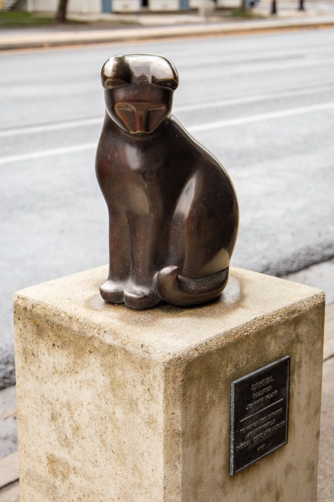 A bronze cat sculpture sitting on a square stone pedestal on a sidewalk.