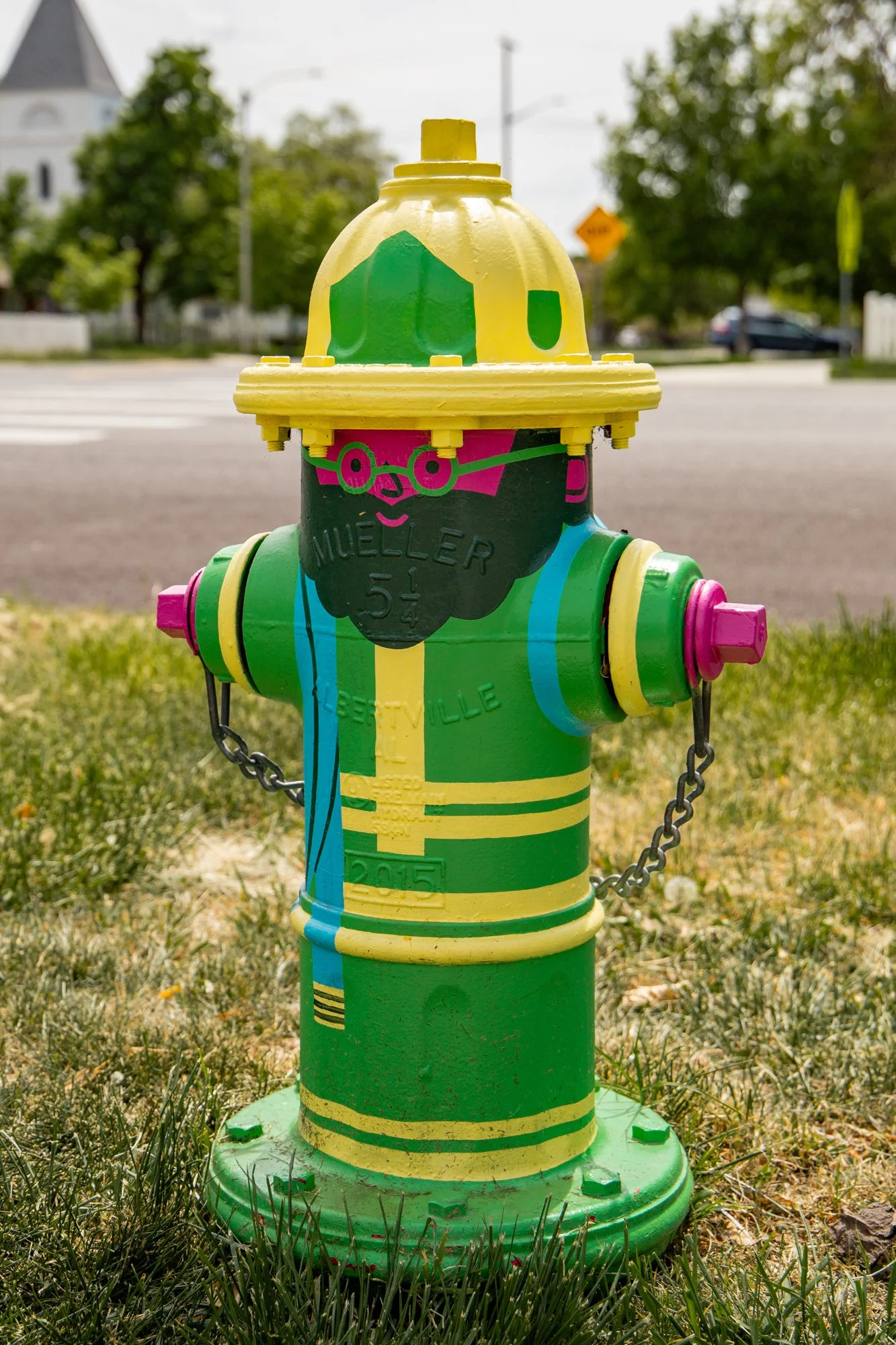 Colorful fire hydrant painted with bright green, yellow, pink, and blue stripes, with a cartoon face and glasses, located on a grassy area near a street with trees and houses in the background.