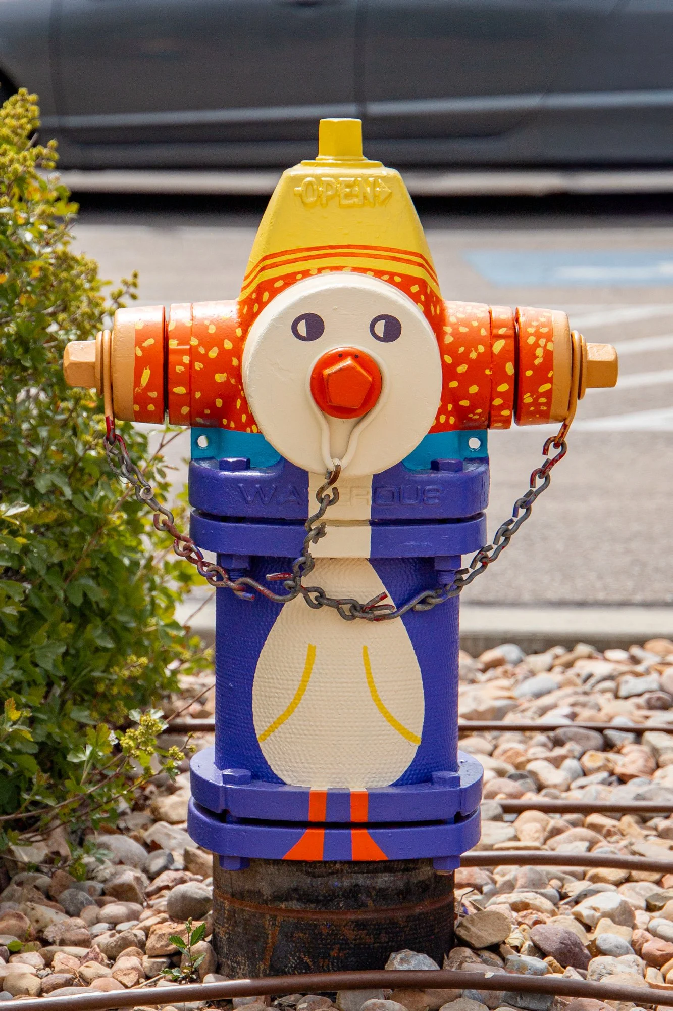 Colorful fire hydrant painted to look like a chicken, with a yellow comb, orange beak, and blue body, situated next to gravel and some green shrubs.