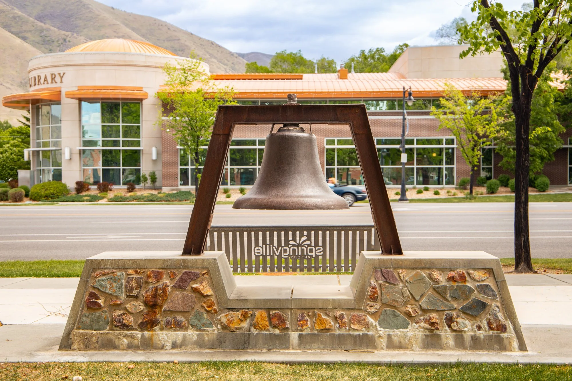 A large bronze bell on a concrete and stone base in front of a library building with large glass windows and trees, with mountains in the background.