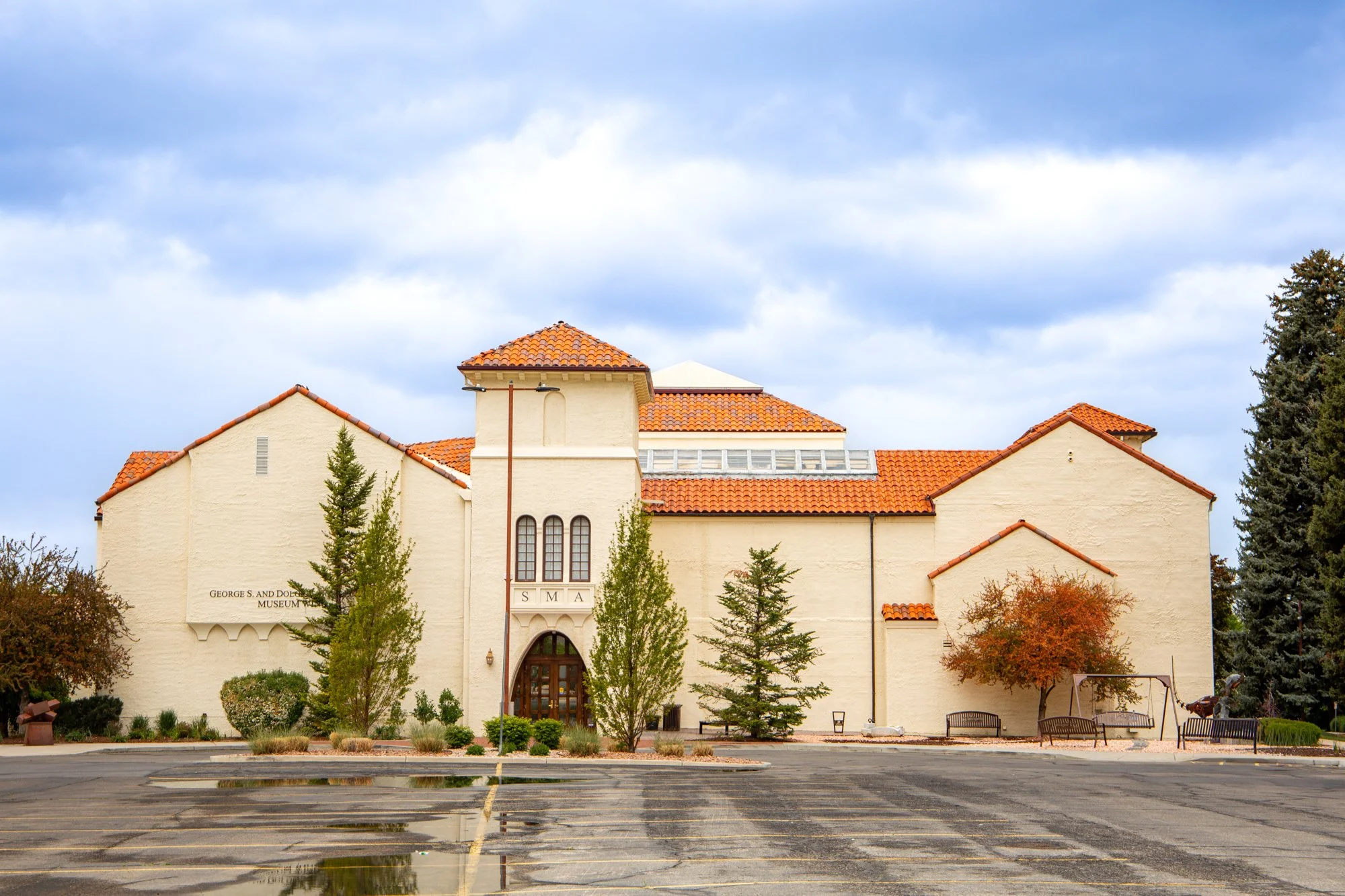 The George S. and Dolores Bishop Museum building with a beige facade and red tile roof, surrounded by trees and a parking lot with a reflective puddle, under a cloudy sky.