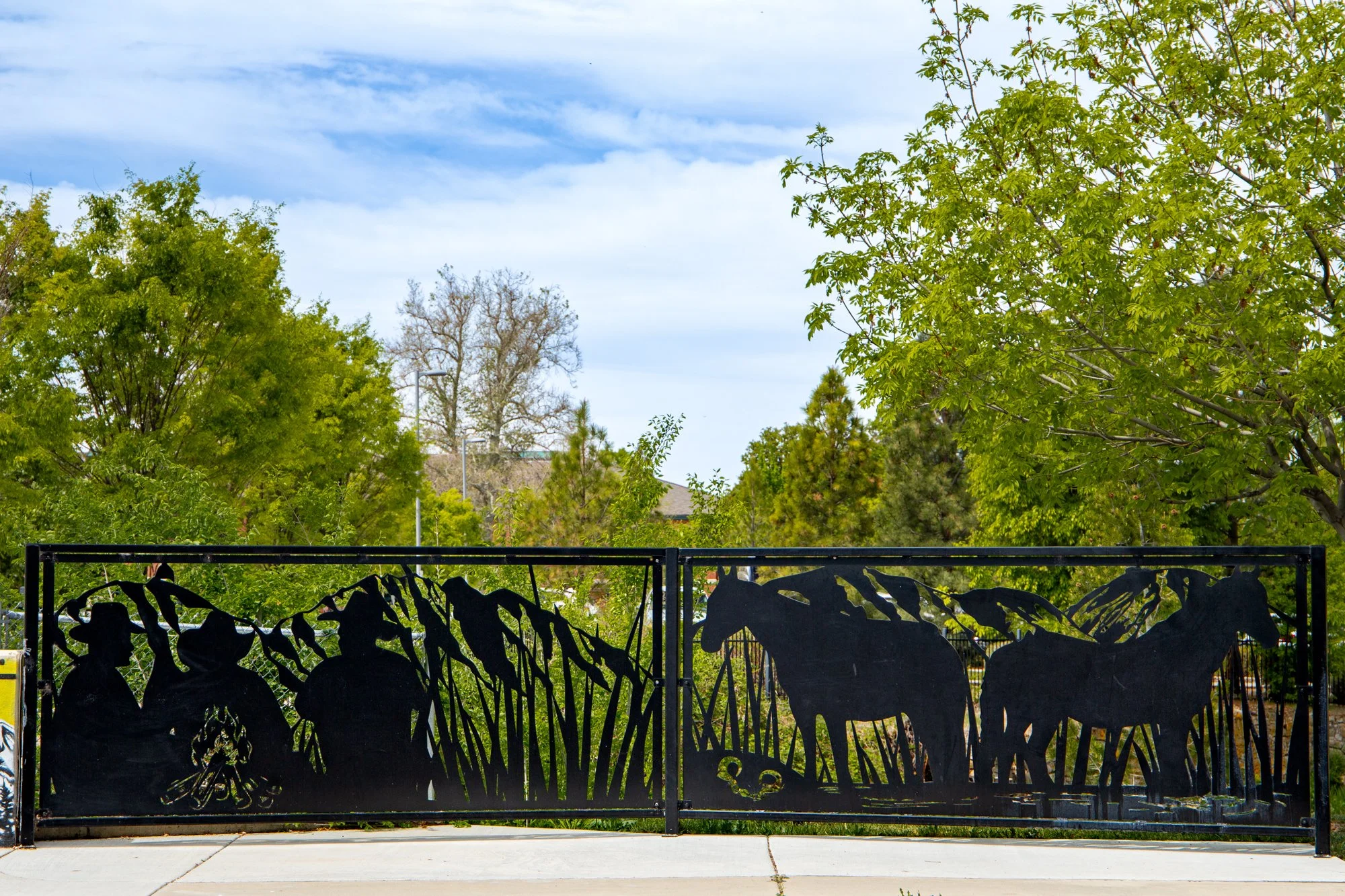 A black metal fence with cutout shapes of animals, including a lion, elephant, and bird, in front of a garden with various trees and green foliage under a partly cloudy sky.