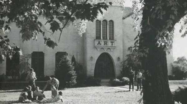 People sitting and standing outside a building with an ART sign above the entrance, surrounded by trees.