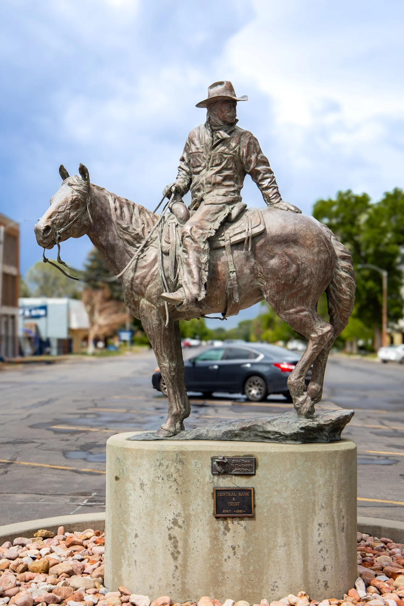 Bronze statue of a man on a horse in a small town setting with buildings and trees in the background, during daytime with cloudy sky.