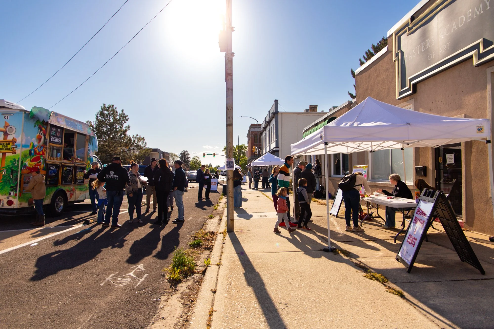 People gather outside on a bright, sunny day at a street fair or community event. There are tents, vendor booths, and a colorful food truck on the left side. Some people are walking, talking, and browsing the booths, while others are seated or working at tables.