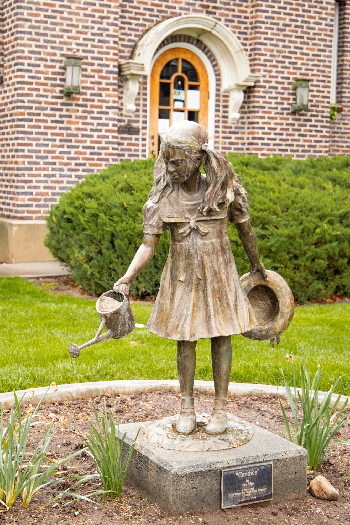 Bronze sculpture of a girl with pigtails holding watering cans, standing on a concrete pedestal with a plaque, located outdoors in front of a brick building with bush landscaping.