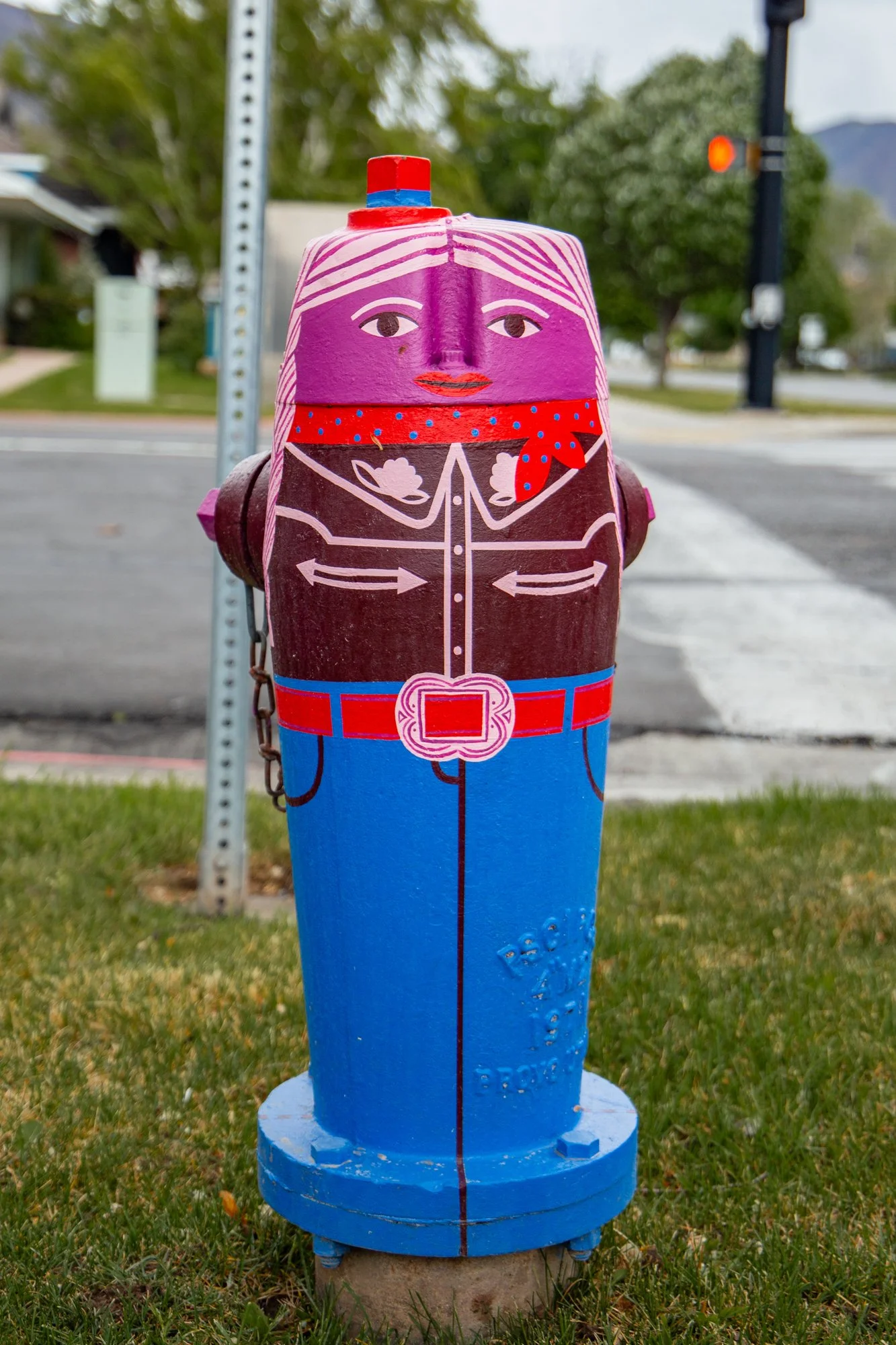 Colorfully painted fire hydrant with a face, wearing a red bandana, located on a grassy area in front of a street with trees and a traffic light in the background.