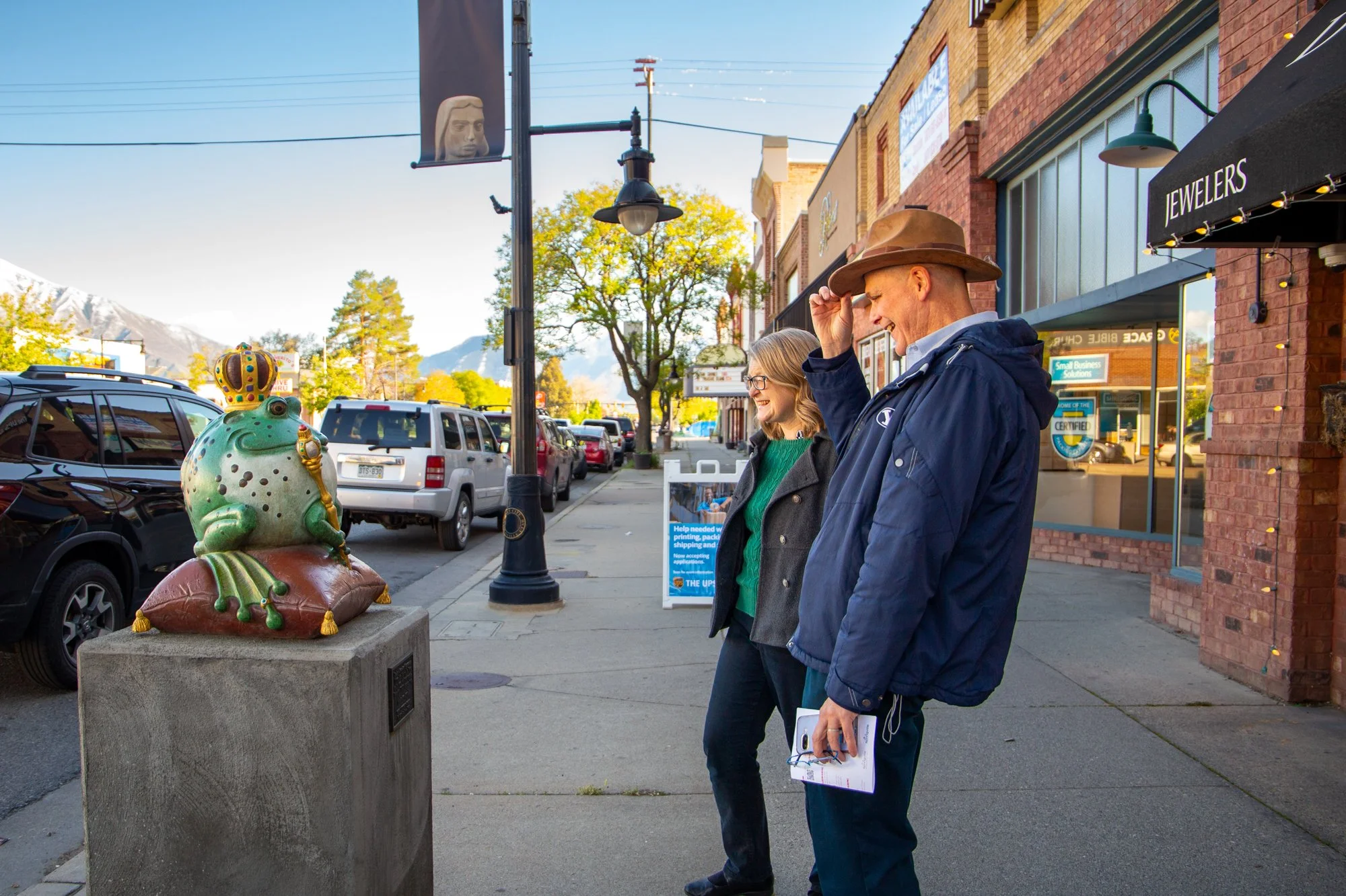 Two people, a woman and a man, standing on a sidewalk in a small town, looking at a frog statue with a crown. The man is wearing a brown hat and a blue jacket. The woman is wearing glasses and a dark jacket. There are parked cars, brick buildings, tr