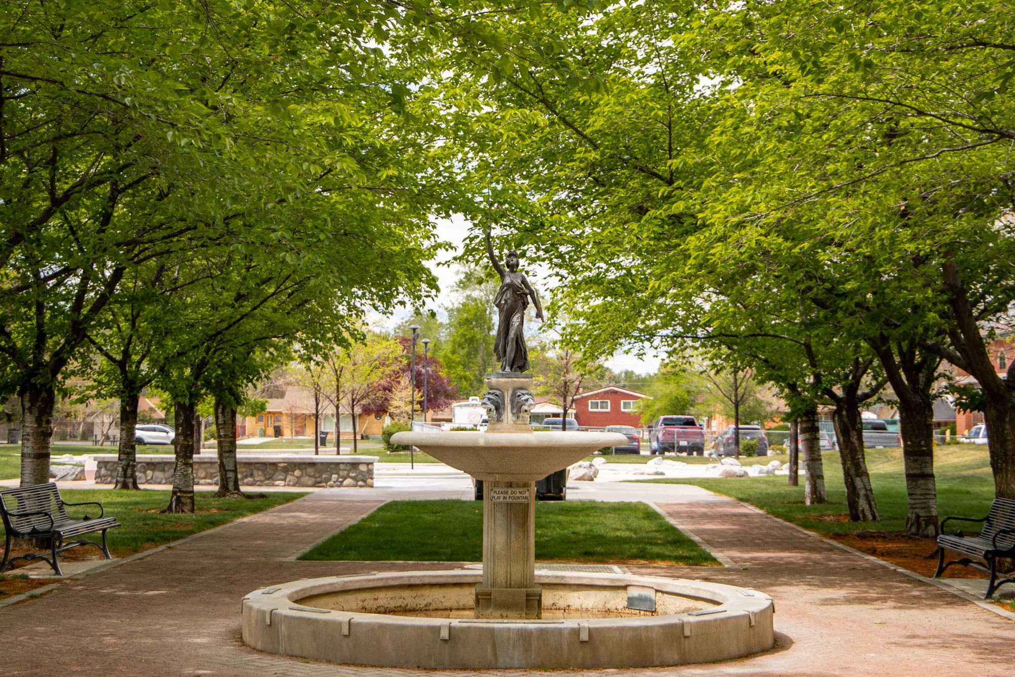 A park with a fountain at the center, surrounded by green trees and benches. In the background, there are houses and parked cars. The fountain features a statue of a woman holding an object, with a sign that warns not to play in the fountain.