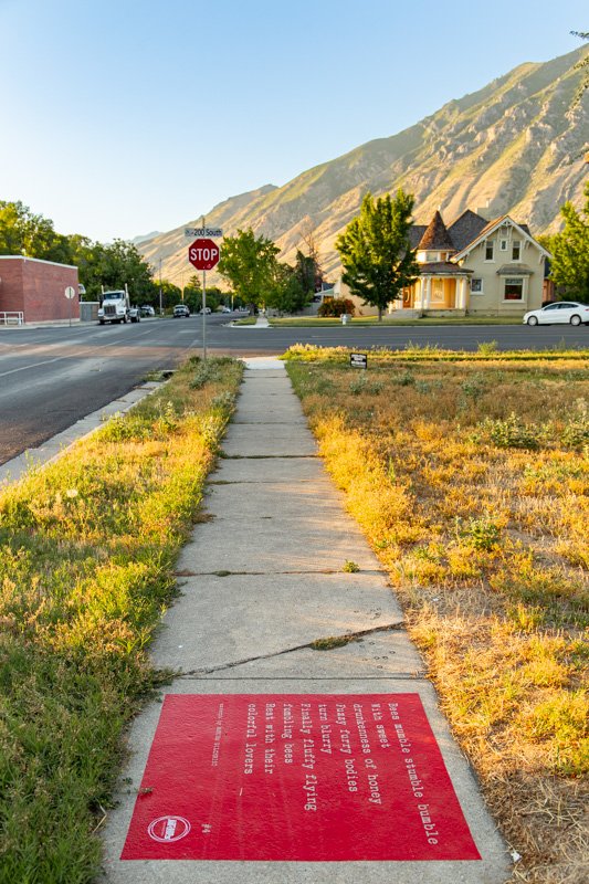 Red sidewalk poem installed along a neighborhood sidewalk with mountain views, a stop sign, and historic homes visible in the distance in Springville.