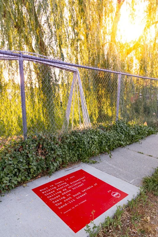 Red sidewalk poem installed beside a sidewalk bordered by ivy and a chain-link fence, with large willow trees overhead in Springville.