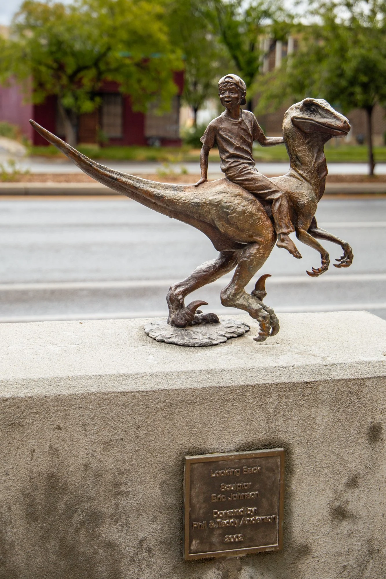 Bronze sculpture of a young boy riding a dinosaur, specifically a small theropod. The sculpture is mounted on a stone base with a plaque, and is located outdoors along a street with trees and buildings in the background.