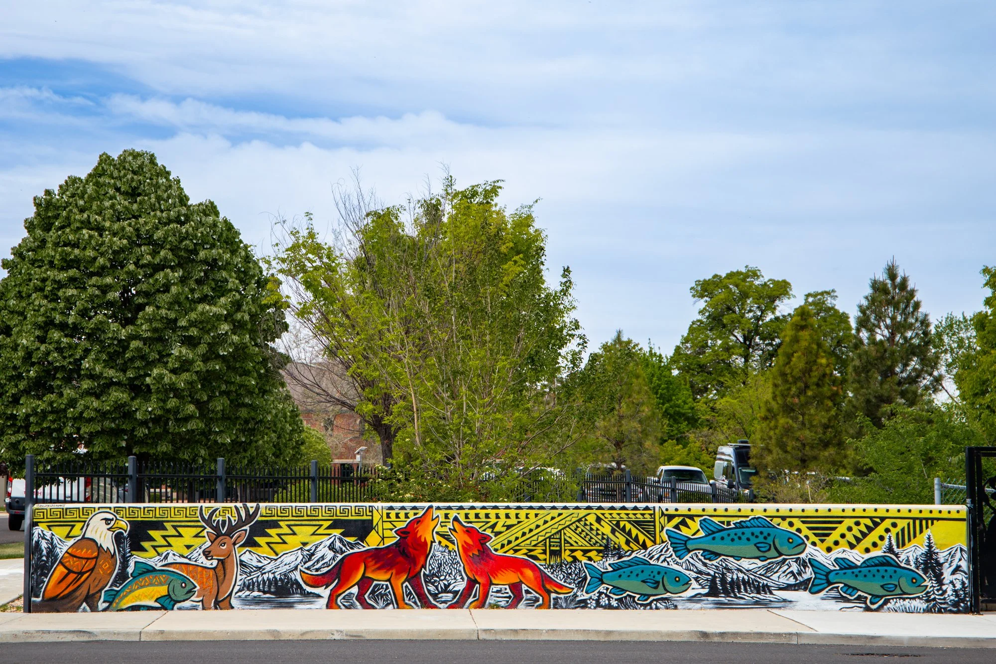 Colorful mural featuring a bald eagle, mountain goats, wolves, a fish, and mountains in the background, with trees and parked cars behind fence