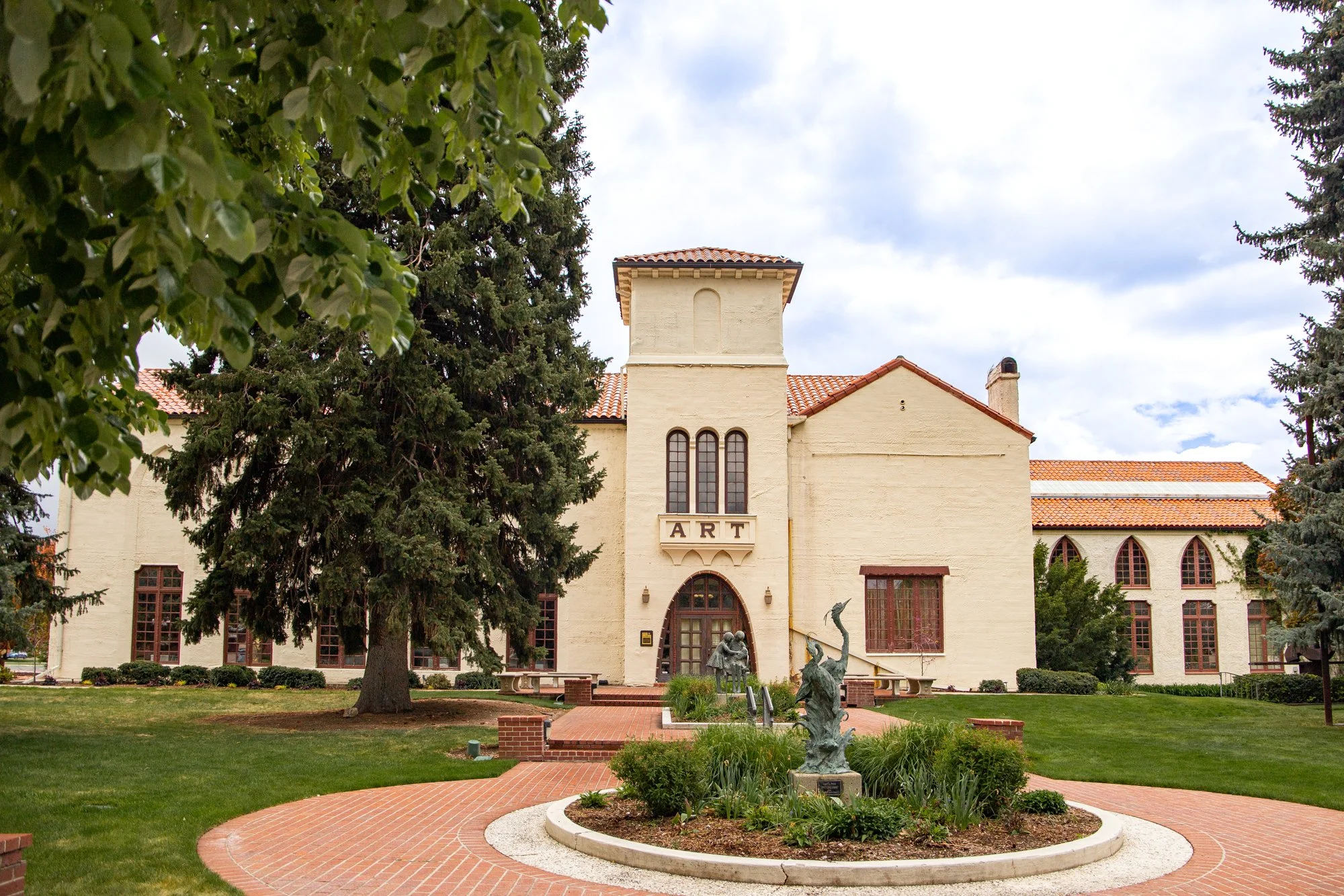 Exterior view of a large, old building with a sign that reads 'ART'. The building has a beige stucco facade, arched windows, and a red-tiled roof. In front, there is a landscaped garden with sculptures, bushes, and a paved walkway. Tall trees surroun