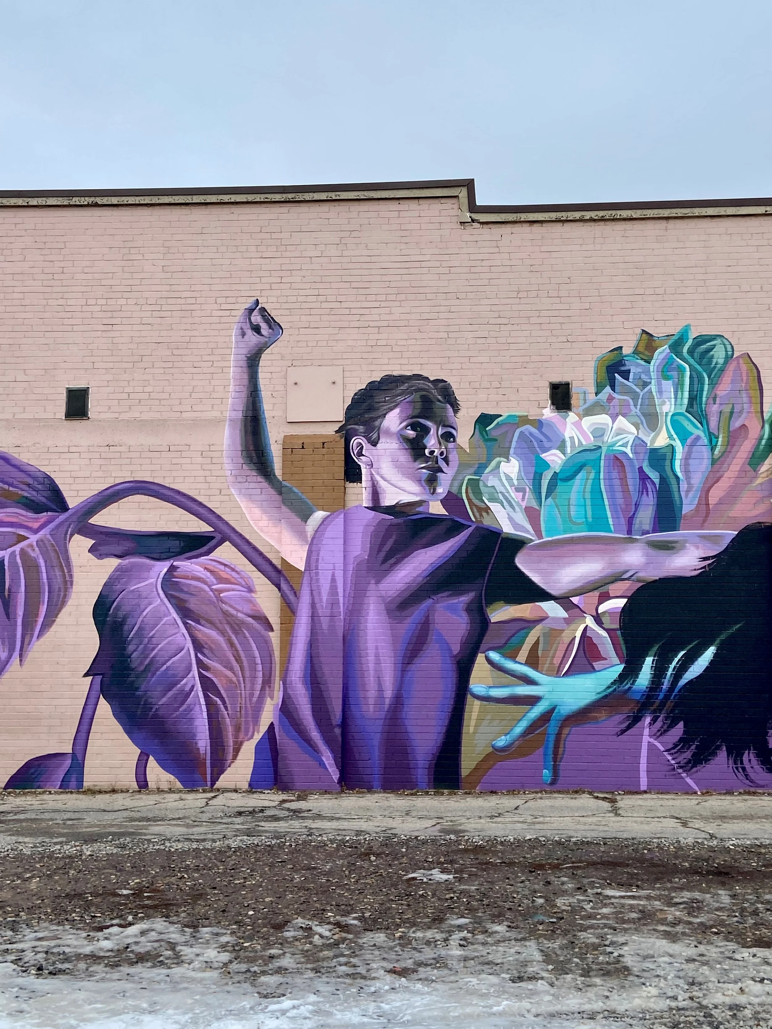 A colorful mural on a brick wall depicting a young woman dancing, surrounded by large purple and teal flowers, with a clear blue sky above.