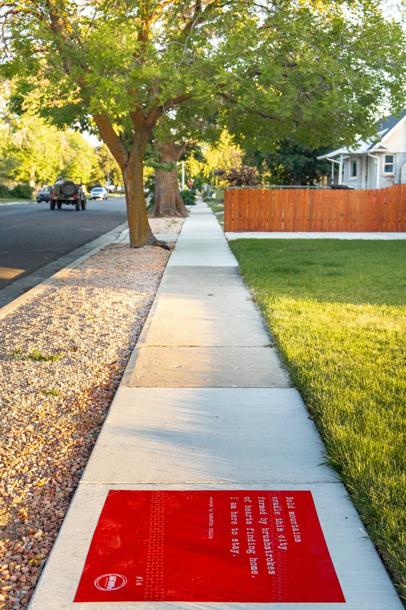 Red sidewalk poem installed along a tree-lined residential sidewalk in Springville, with gravel landscaping, green lawns, and neighborhood homes in the background.
