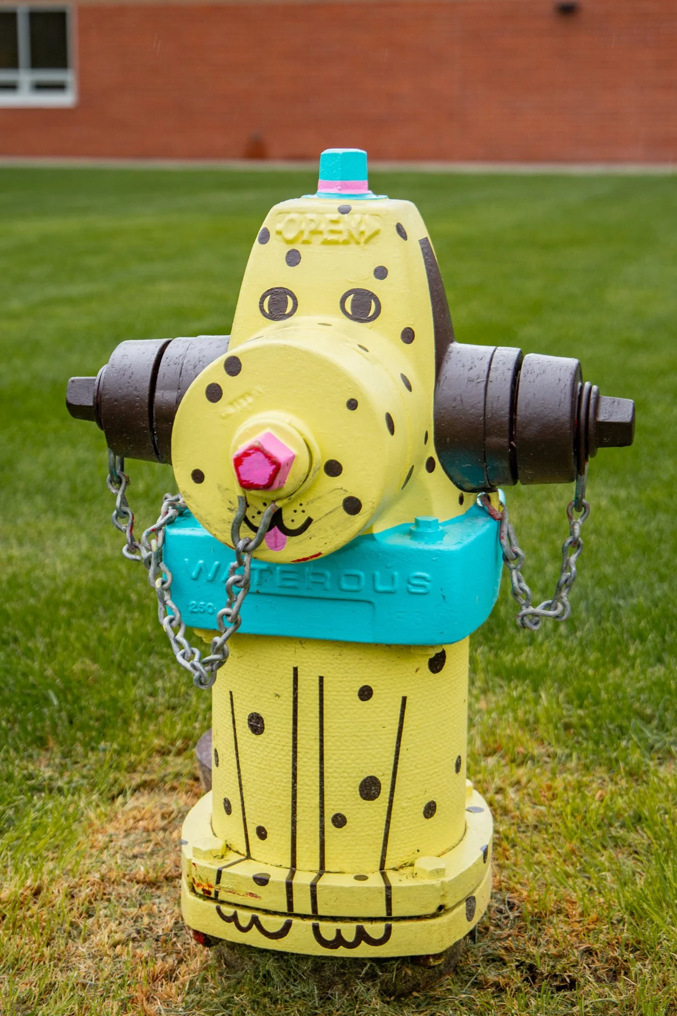 Colorful fire hydrant decorated like a yellow giraffe with black spots, cartoonish face, and a red nose, set on a grassy outdoor area with a brick building in the background.