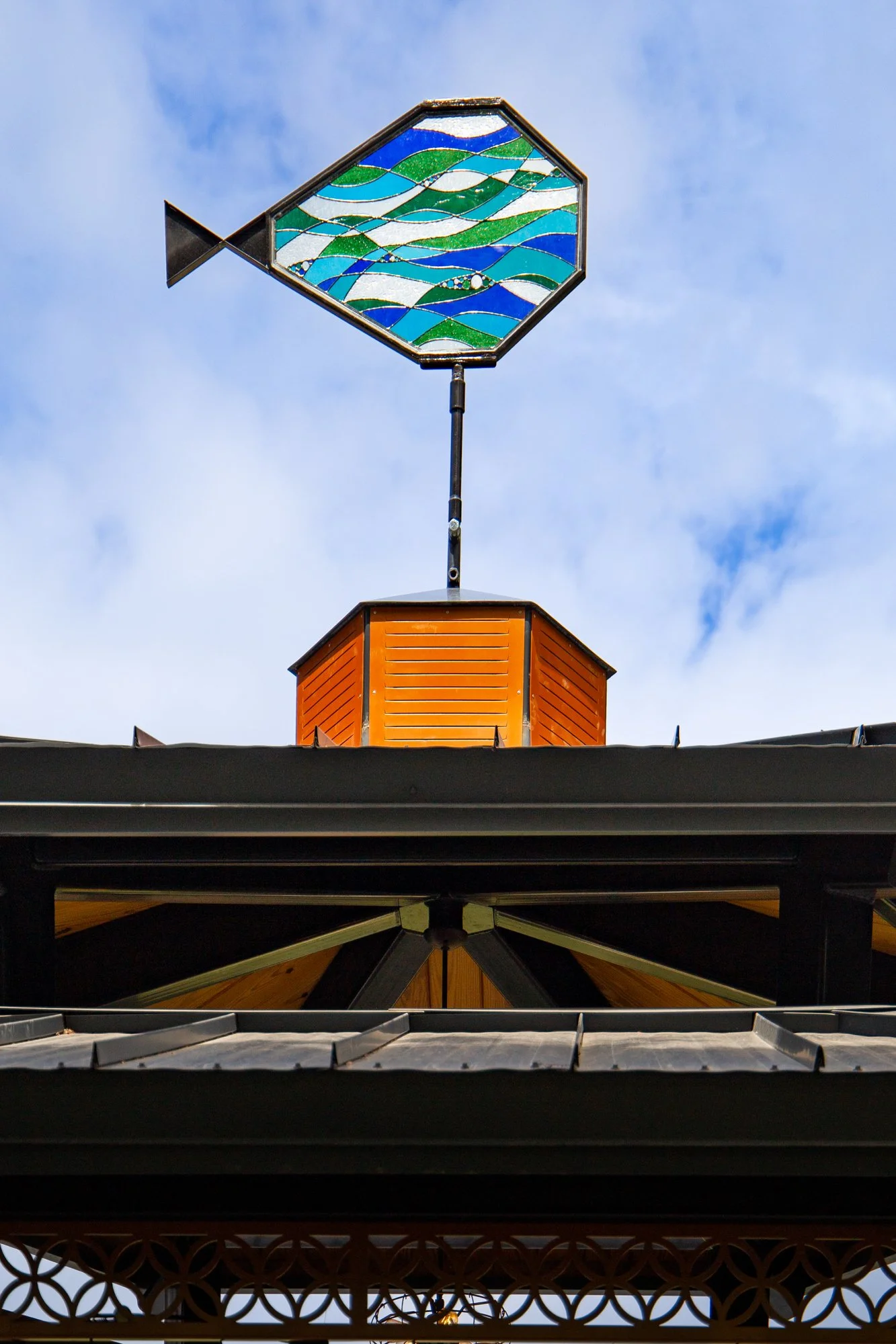 A decorative orange vent hood with a stained glass fish design on top, set against a blue sky with some clouds.