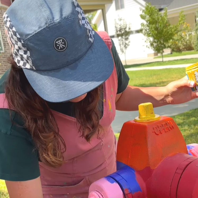 A person wearing a blue and checkered cap, a pink jumpsuit, and a dark t-shirt, spray painting a fire hydrant outdoors on a sunny day with trees and a house in the background.