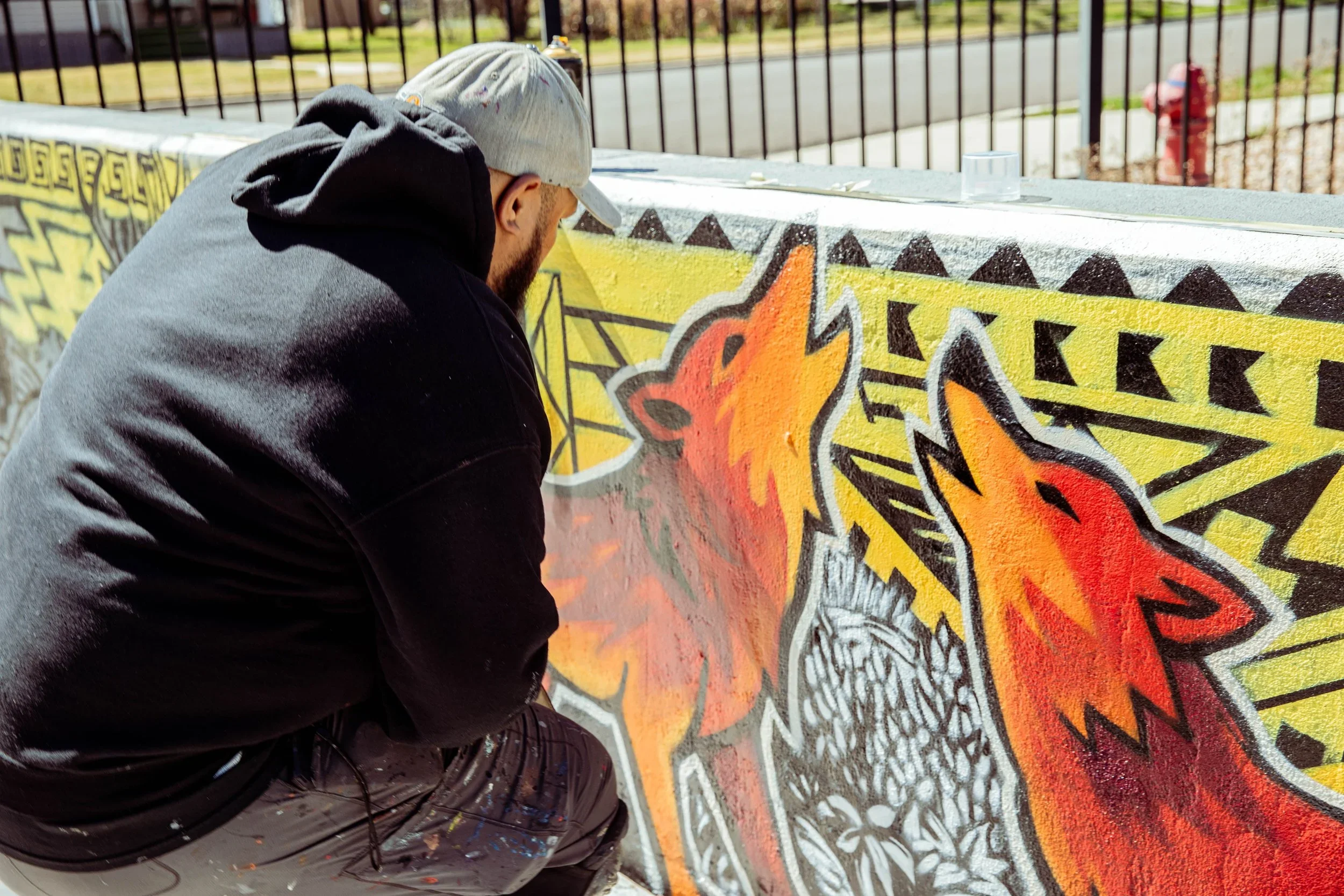 A person with a gray cap and black hoodie spray painting a colorful mural on a concrete wall.