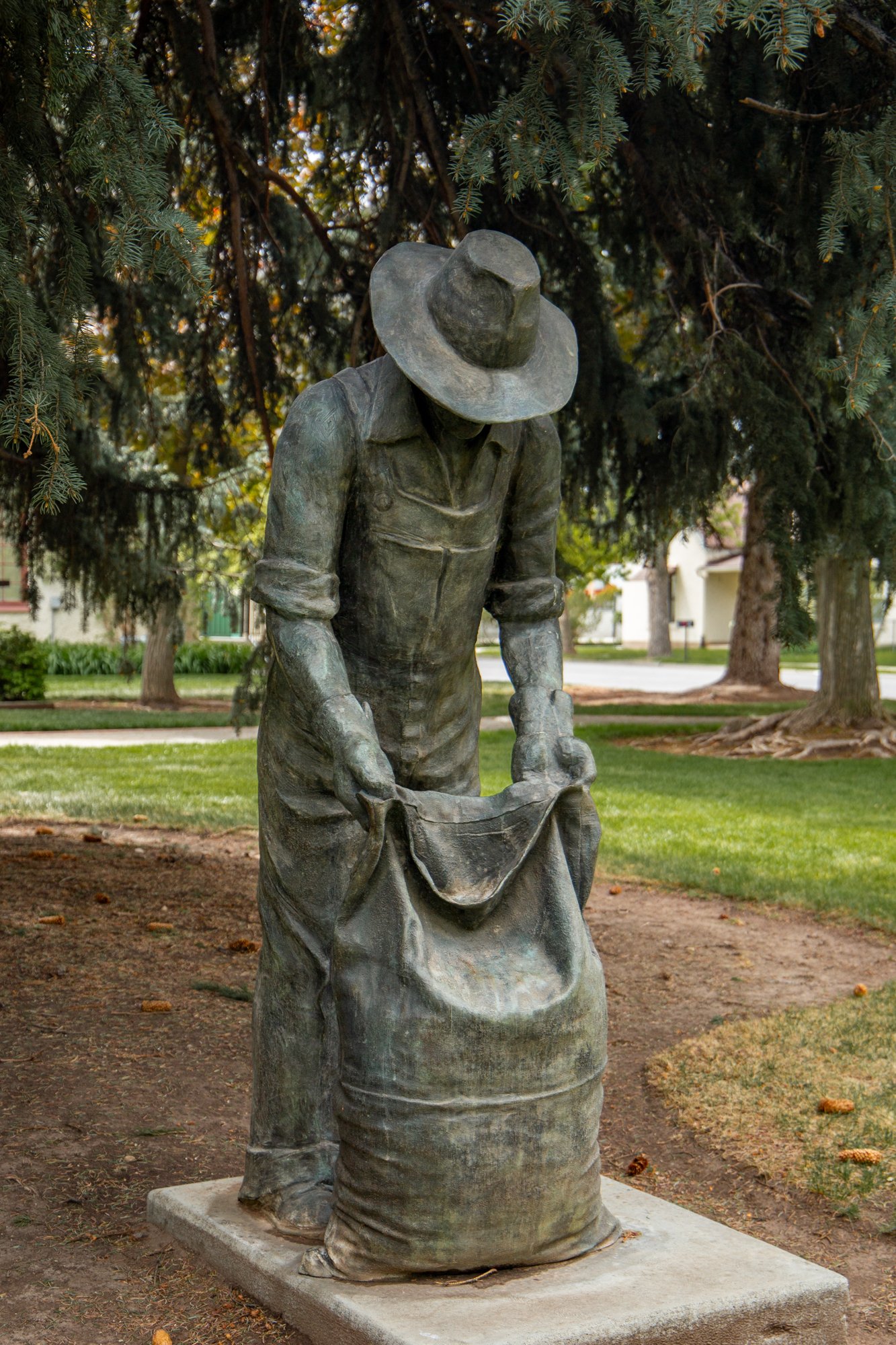 Bronze statue of a woman wearing a wide-brimmed hat, standing outdoors on a concrete base surrounded by trees and grass.