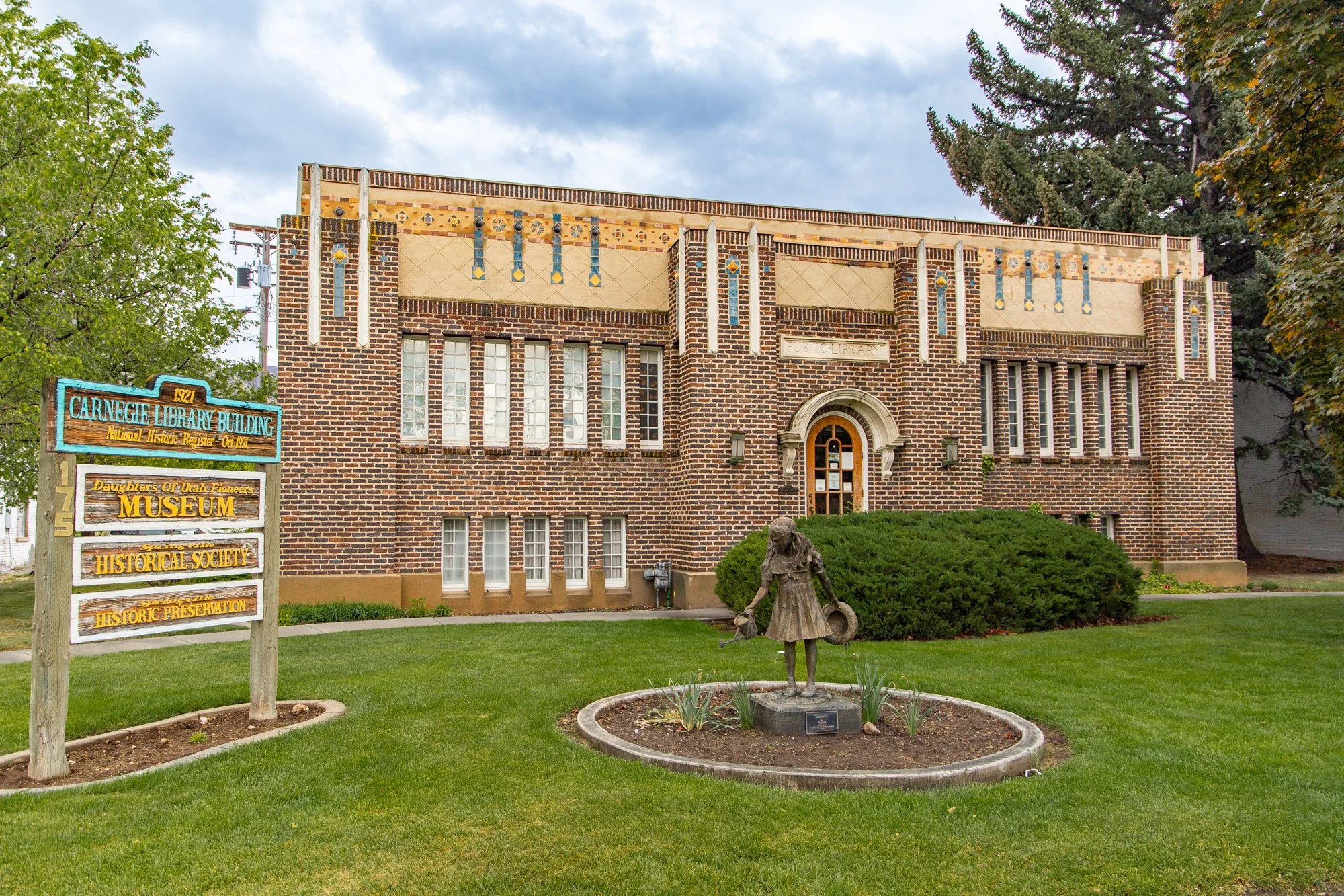 Photo of the Carnegie Library building with brick architecture, decorative tile accents, and a rounded arched door. In front, there's a green lawn, a statue of a girl with a dish, and a sign indicating it is a museum and part of the Utah Pioneers.