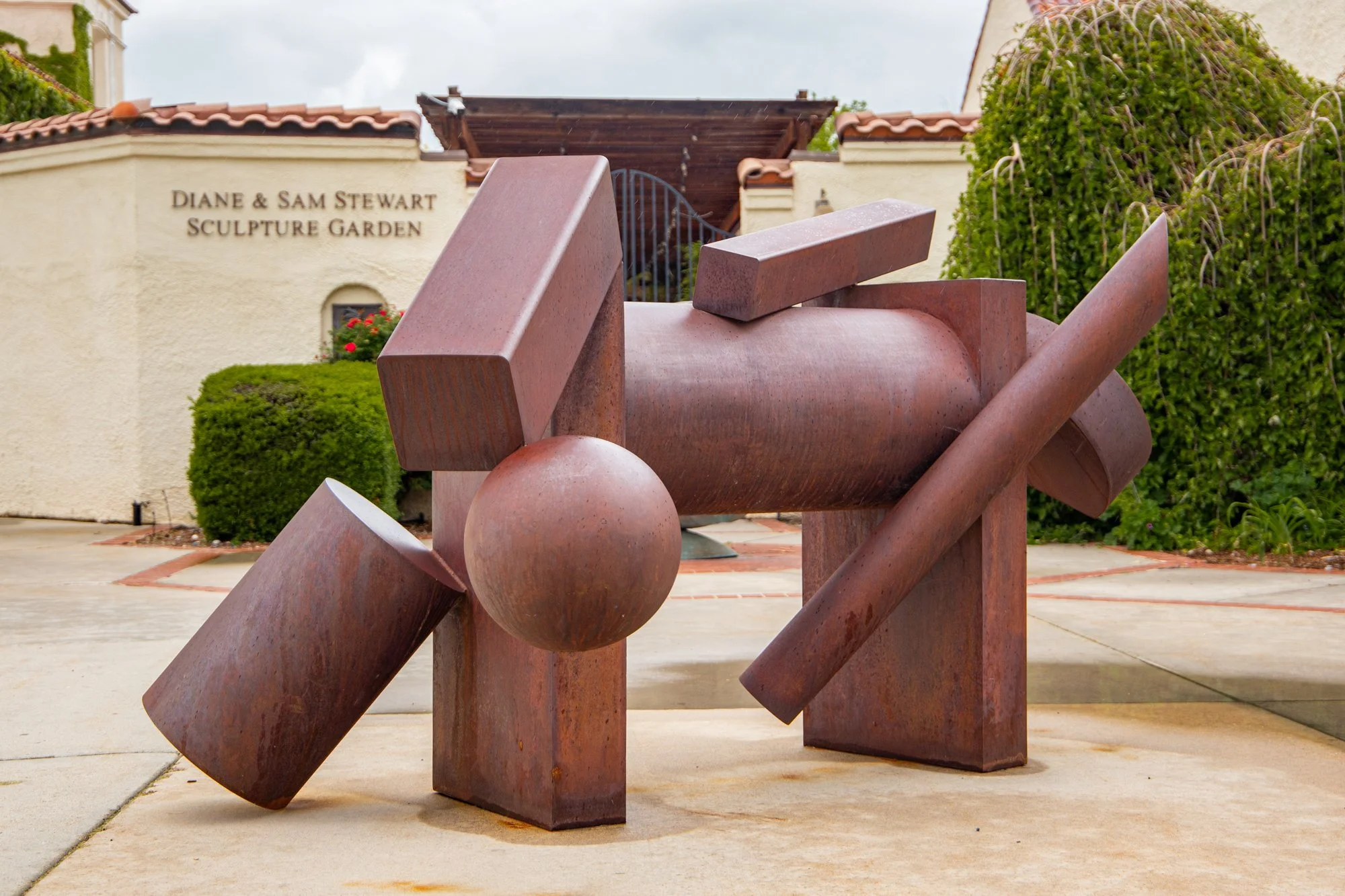 Abstract sculpture made of pinkish-brown stone with geometric shapes, including a sphere, rectangles, and slabs, placed outdoors near greenery and a building with the sign 'Diane & Sam Stewart Sculpture Garden'.
