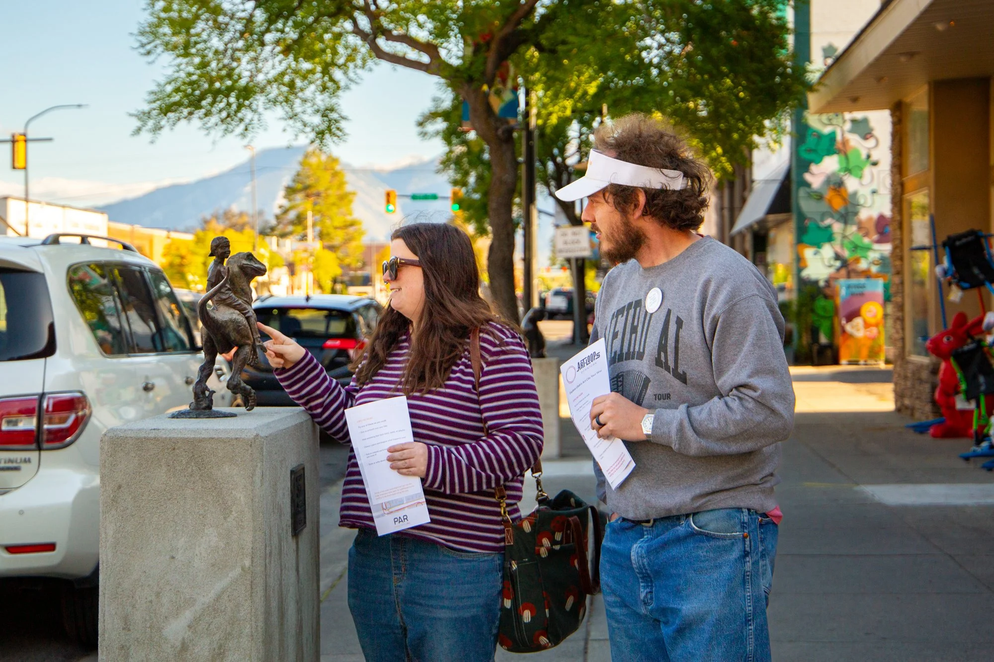 A woman and a man standing on a sidewalk, looking at a small bronze statue on a pedestal. The woman is smiling and wearing sunglasses, a striped purple and white sweater, with a bag on her shoulder and holding a brochure. The man is smiling, wearing a gray sweatshirt, a white visor, and holding a brochure. There are parked cars, trees, and storefronts in the background on a sunny day.