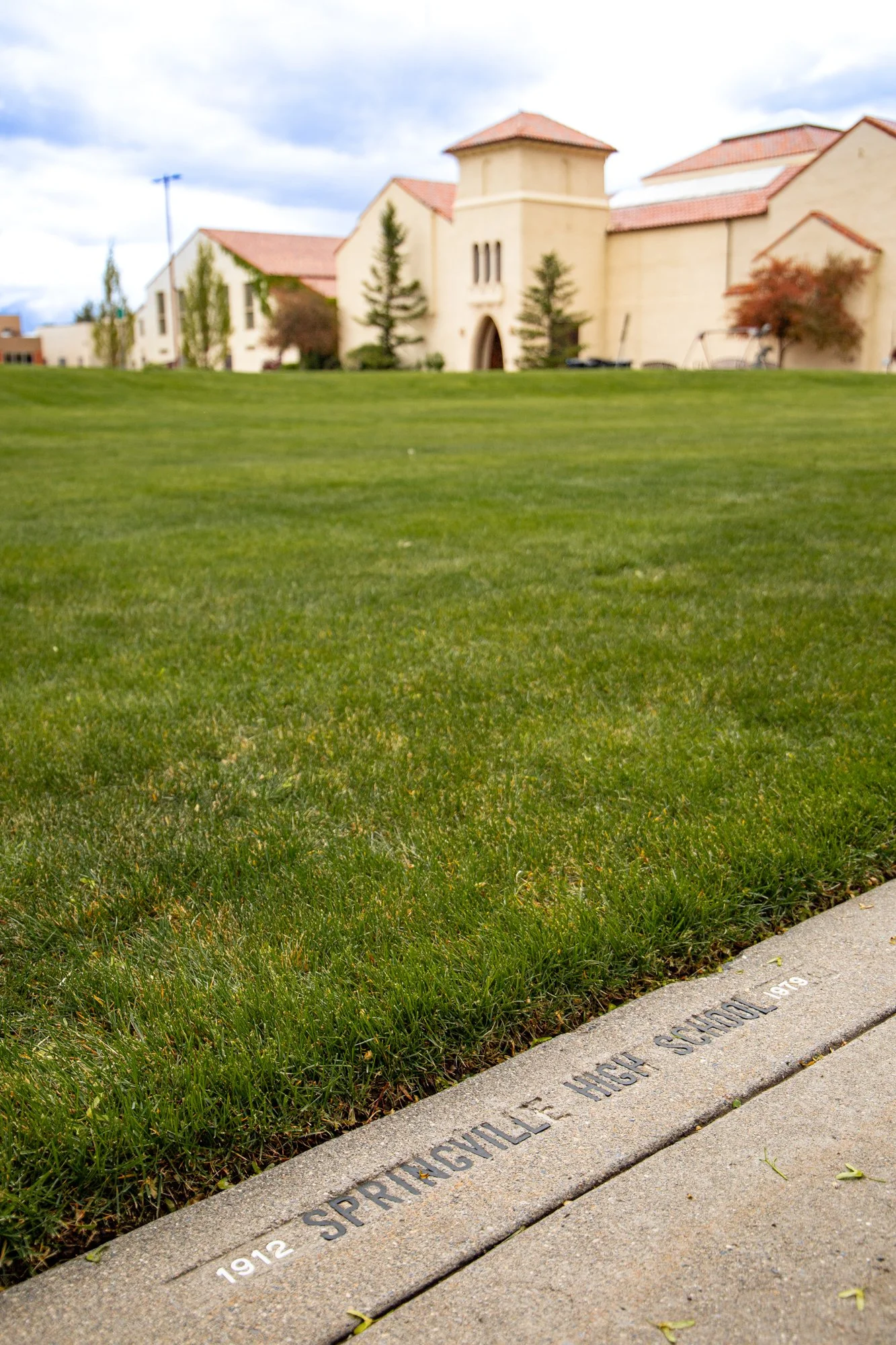 Close-up of a sidewalk engraved with the text "1912 SPRINGVILLE HIGH SCHOOL" next to a grassy area, with a large building with beige walls and red-tiled roof in the background under cloudy sky.