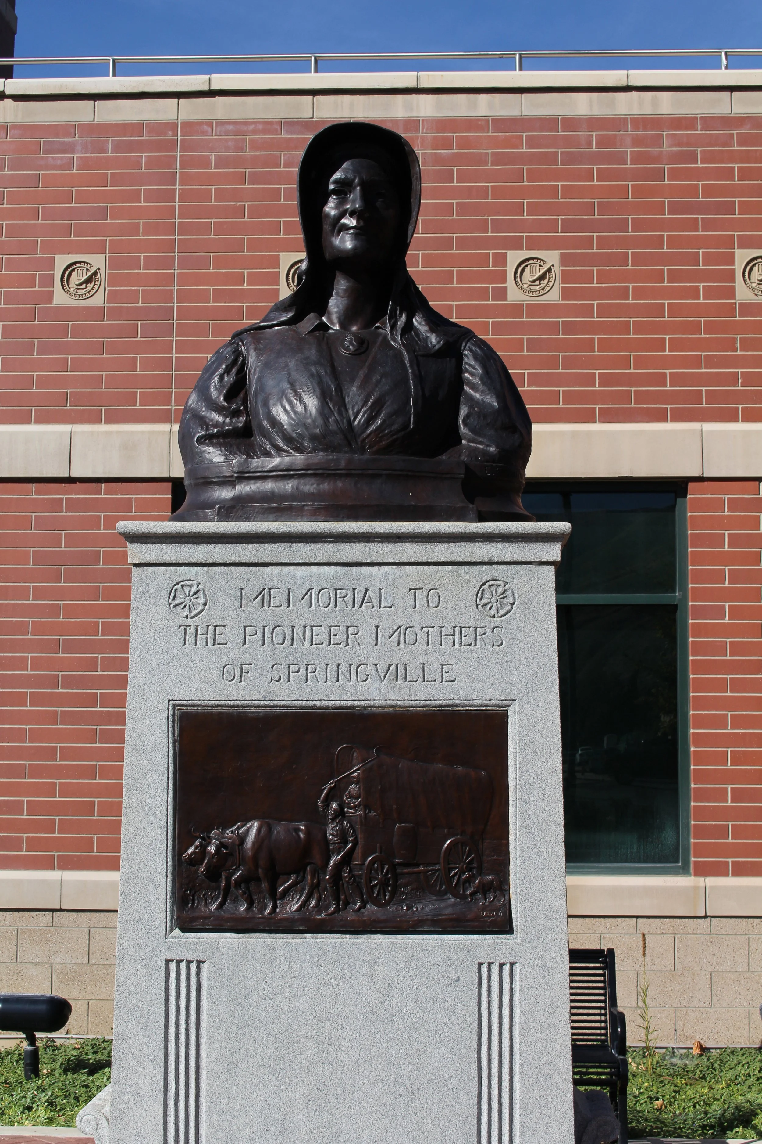 Bronze bust of a woman on a granite pedestal in front of a brick building with a blue sky overhead. The pedestal has an inscription that reads, 'A memorial to the Pioneer Mothers of Springville' and a bronze relief depicting early settlers and a covered wagon.