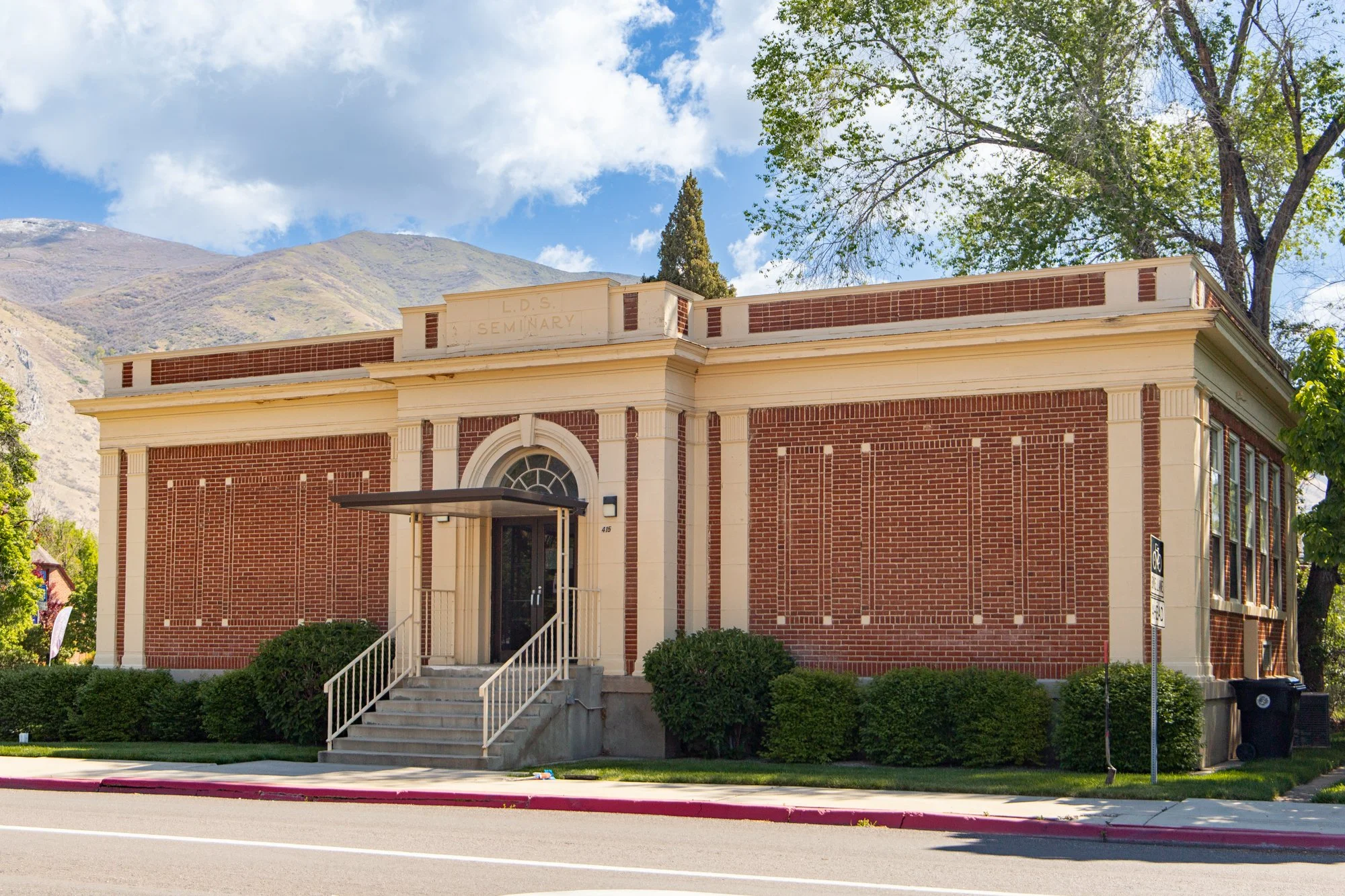 Front view of a historic brick building labeled LDS Seminary with stairs leading to the entrance, trees surrounding the building, mountain range in the background, and a partly cloudy sky.
