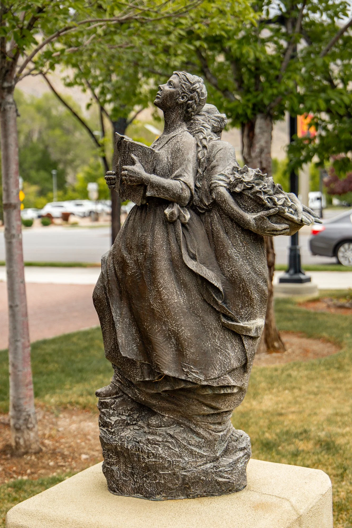 Statue of a woman holding a book, with outdoors and trees in the background.