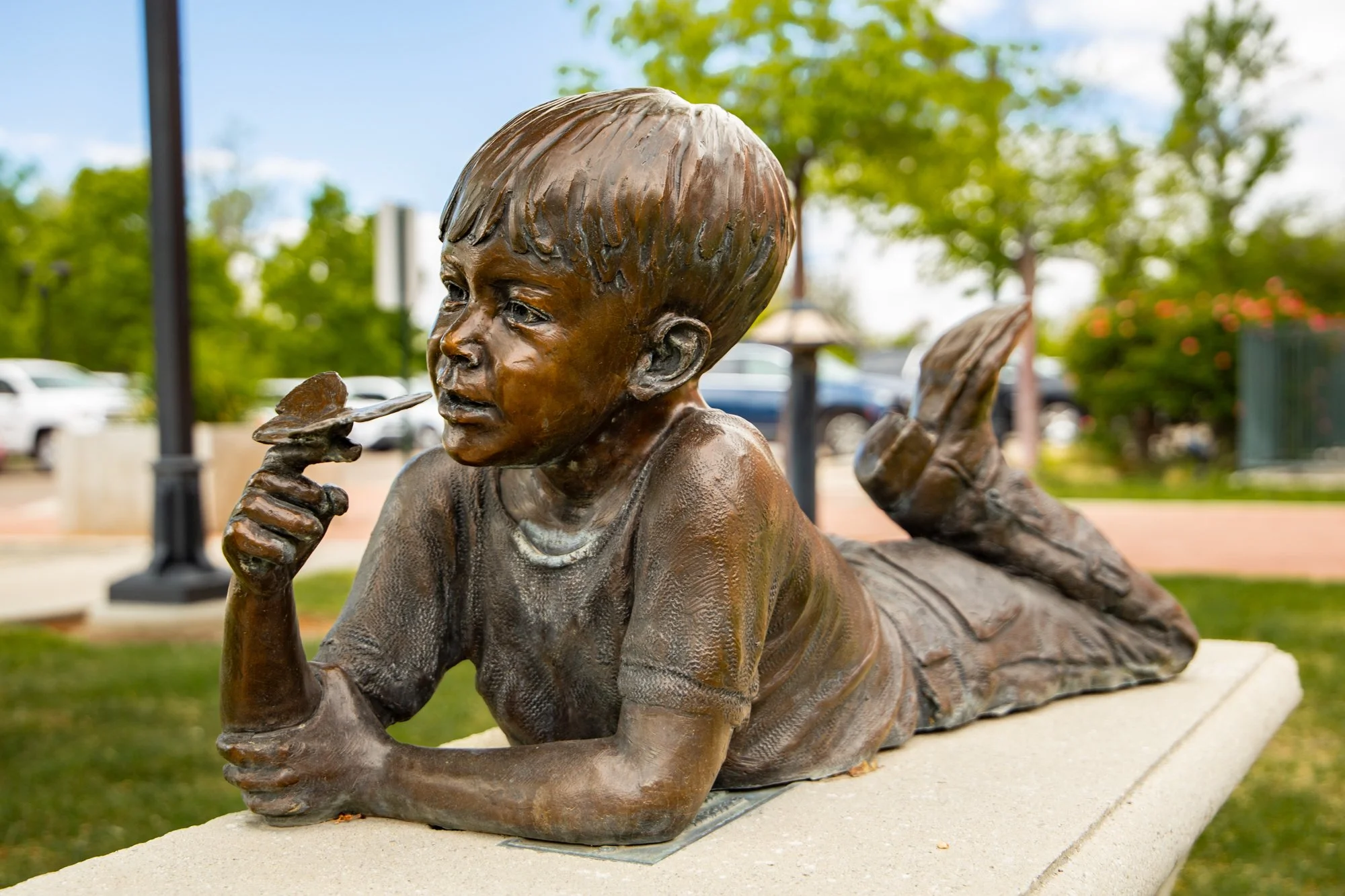 Bronze sculpture of a young boy with short hair lying on his stomach, holding a butterfly in his right hand near his mouth, outdoors with trees, cars, and a gazebo in the background.