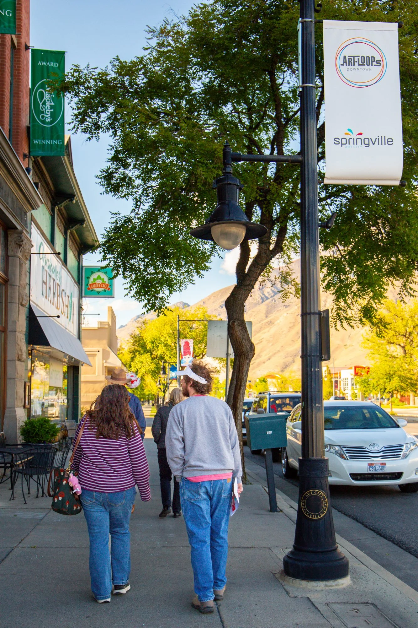 People walking down a city sidewalk lined with shops, street lamps, and trees, with banners hanging from the streetlight pole that read 'Art Tours Downtown' and 'Springville.' Mountains are visible in the background.